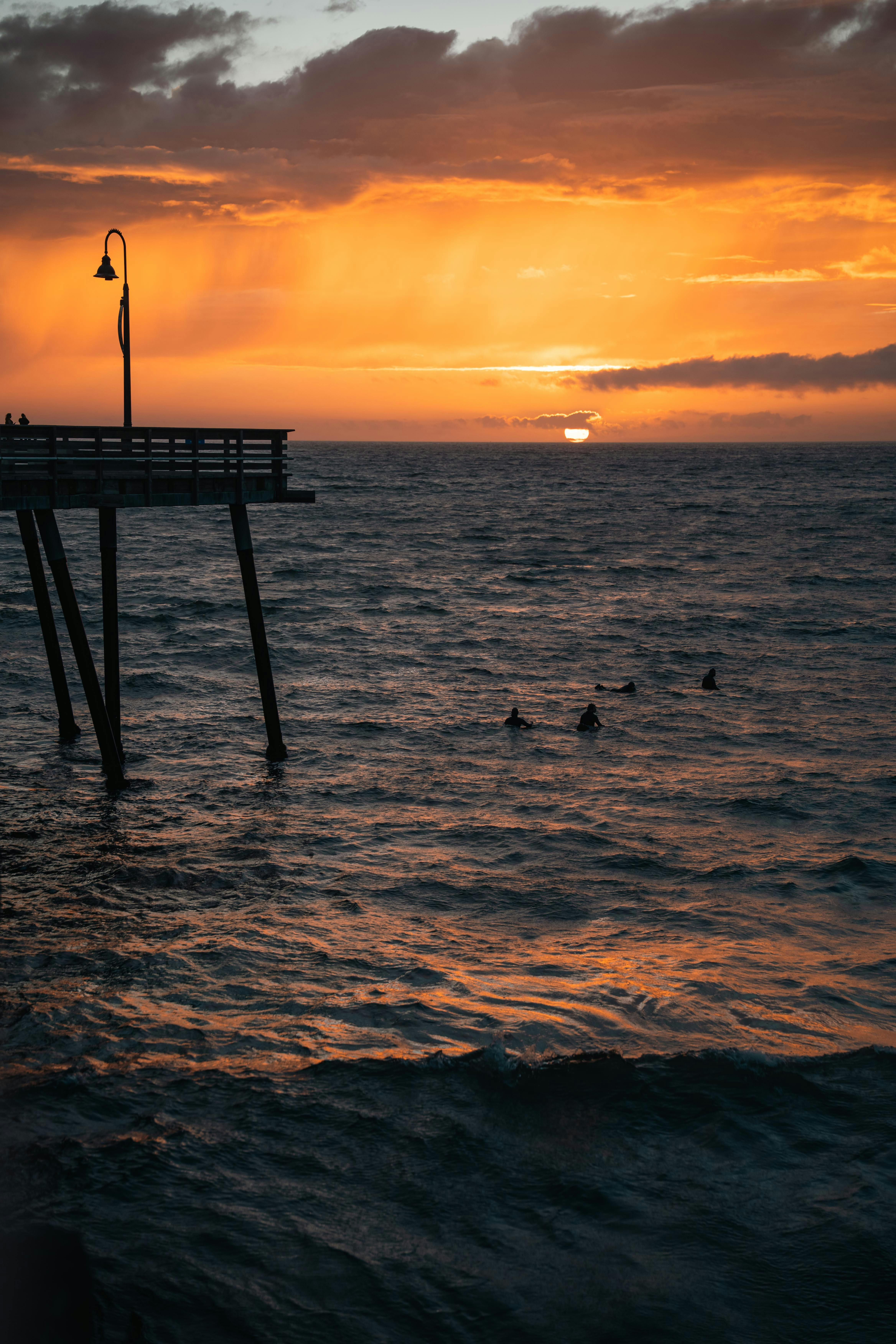 Sunset view with silhouettes of a pier and swimmers in the ocean.