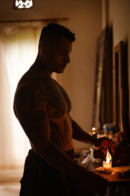 Silhouette of a muscular tattooed man lighting a candle indoors, creating a warm ambiance.