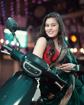 A woman in a saree poses with a smile while sitting on a motorcycle, celebrating indoors.