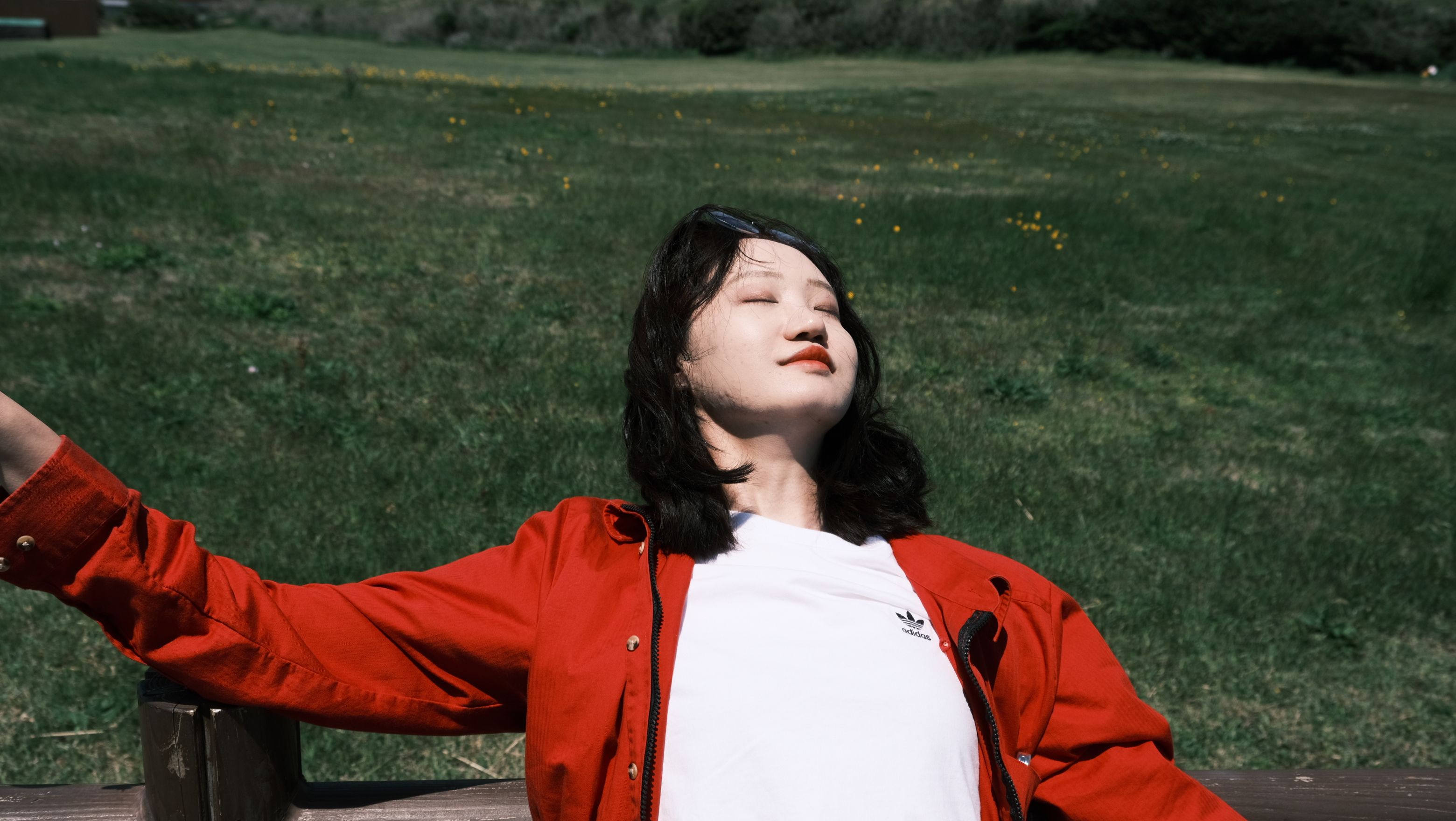 Woman enjoying sunlight outdoors in a park, wearing a red jacket.
