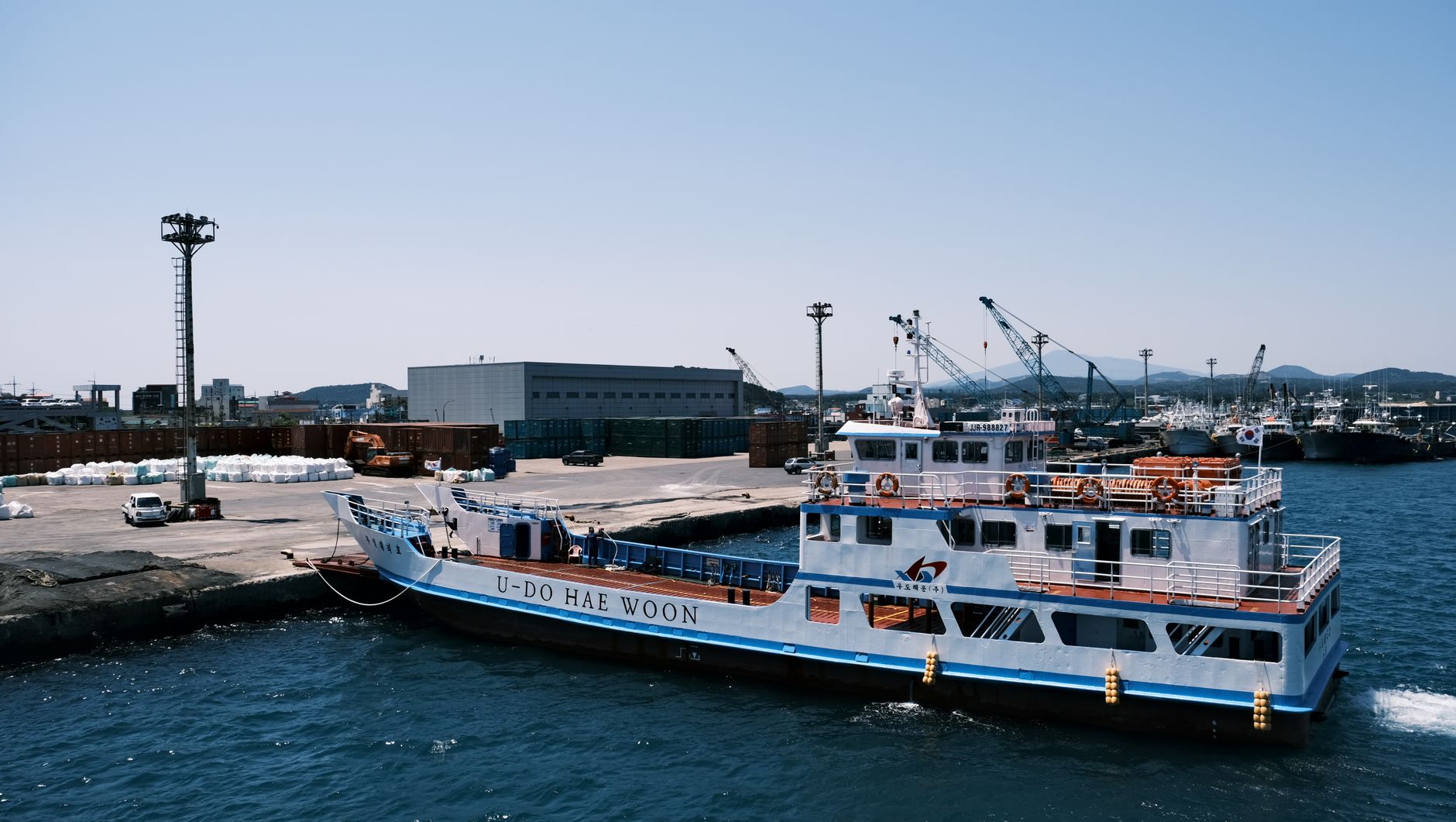 A ferry boat named 'U-Do Hae Woon' docked at a busy industrial harbor with cargo facilities.