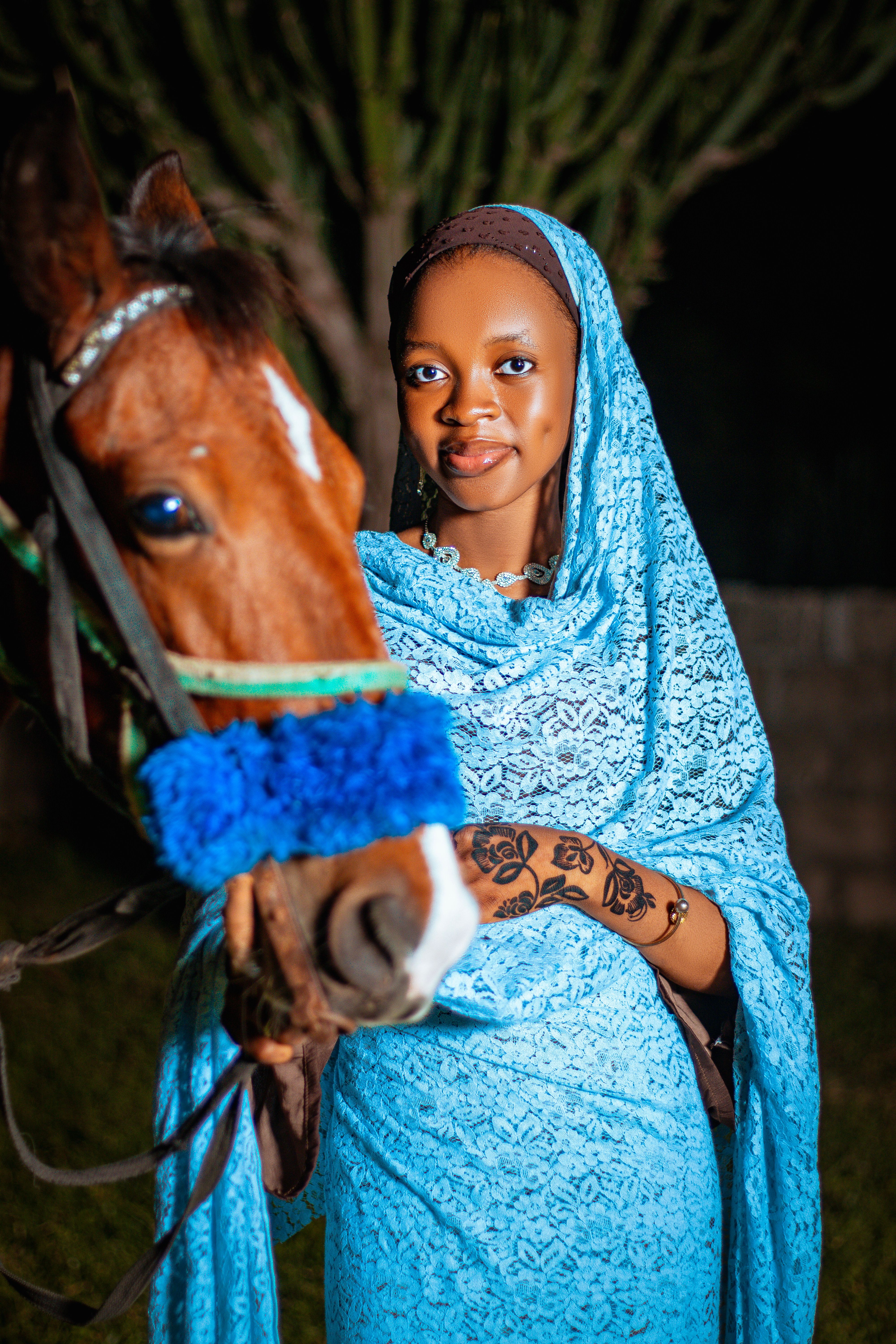 A woman in blue traditional attire poses gracefully with a horse at night.