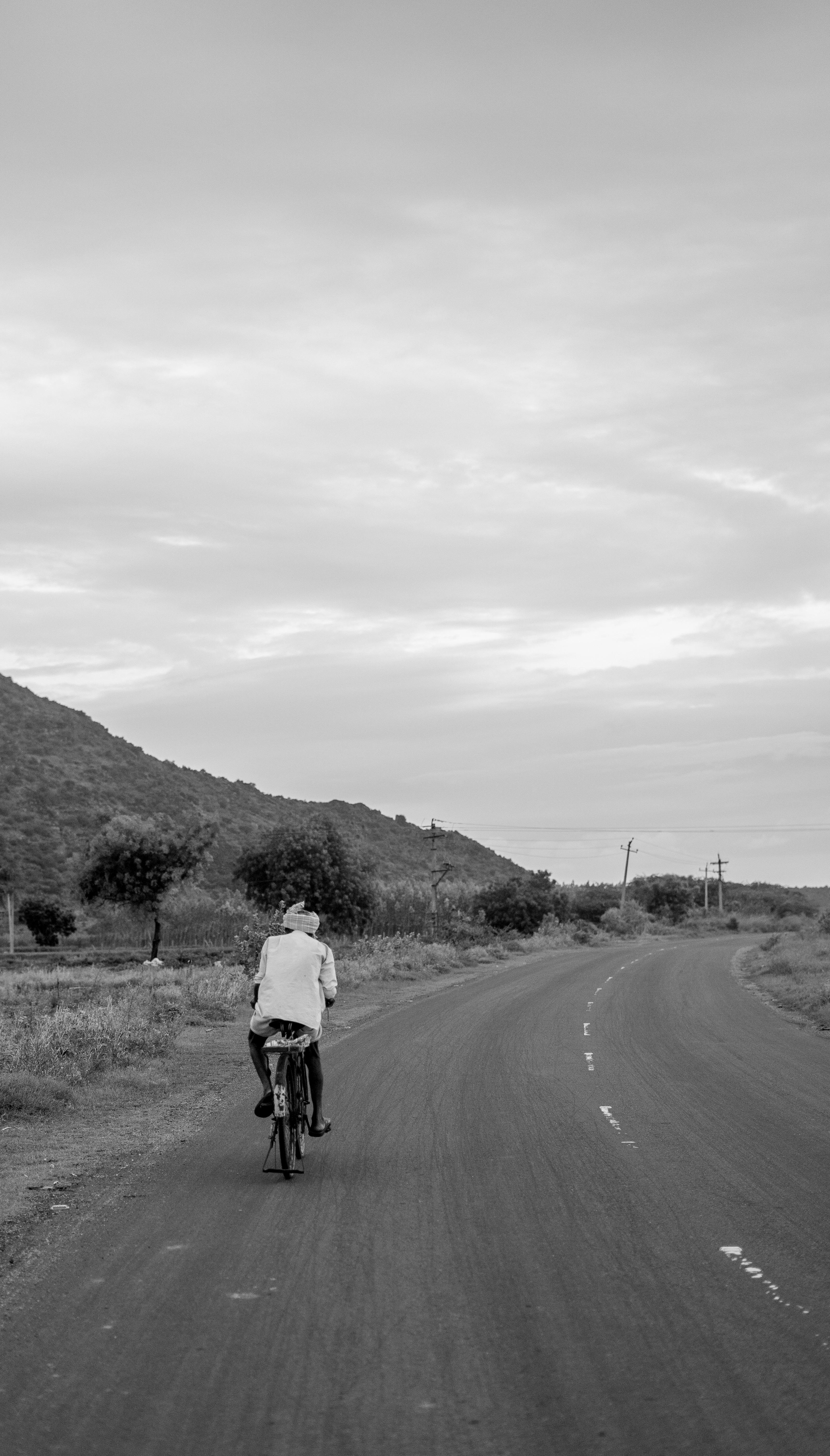 A lone cyclist rides through scenic rural India, surrounded by hills and open road during the day.