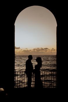 Silhouette of a couple standing near an archway by the sea at sunset, creating a romantic and serene mood.