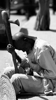 A contemplative elderly man sitting outdoors holding a stick.