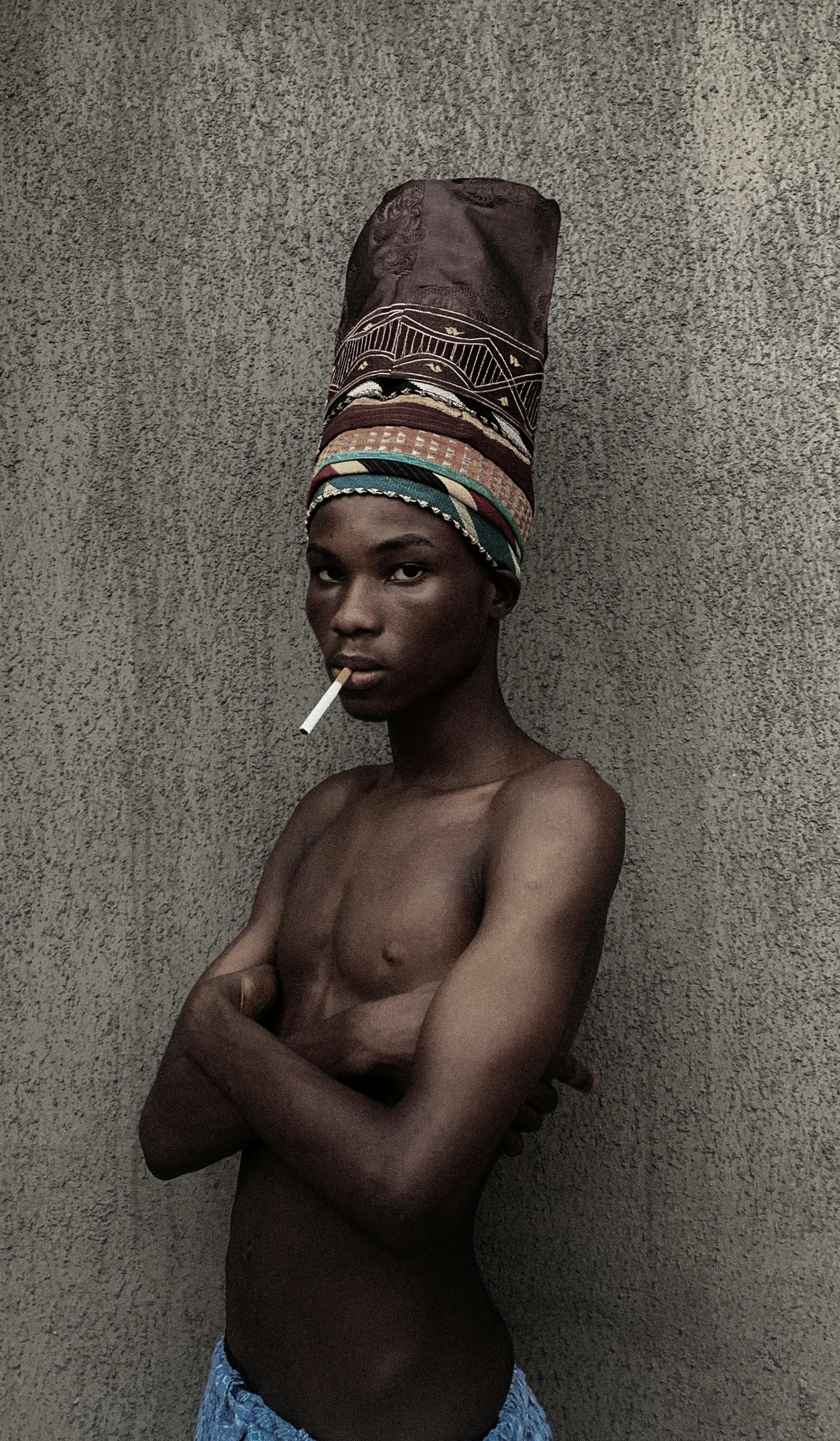 Portrait of a young man wearing a colorful headwrap, holding a cigarette. Calm expression.