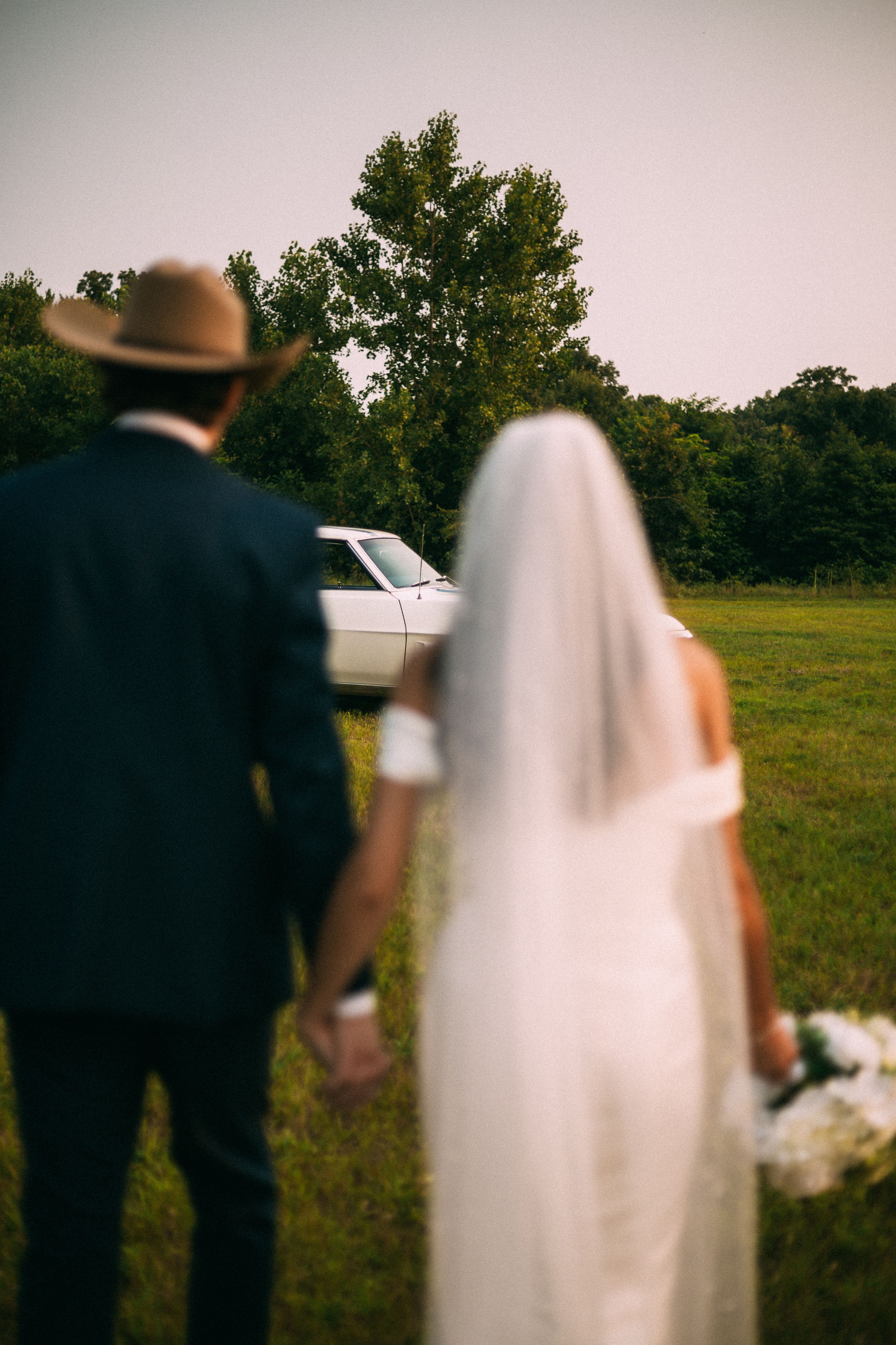 Wedding couple walking towards classic car in picturesque field.