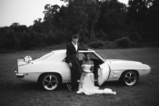 Black and white photo of a couple with a classic car, capturing elegance and vintage style.