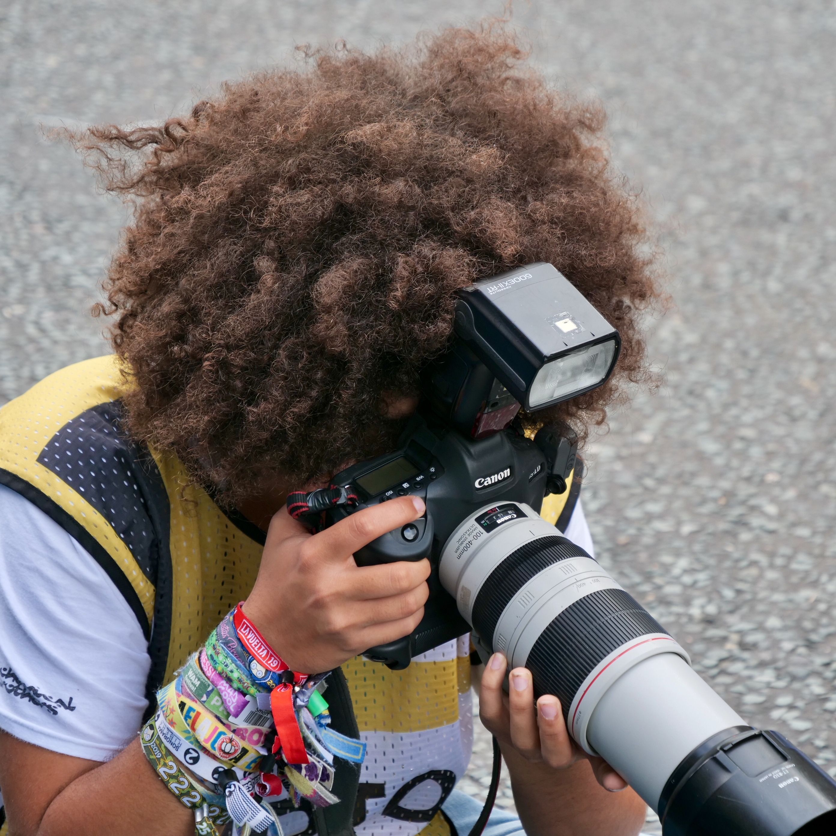 A photographer with distinctive big hair captures images using a camera with a telephoto lens outdoors.