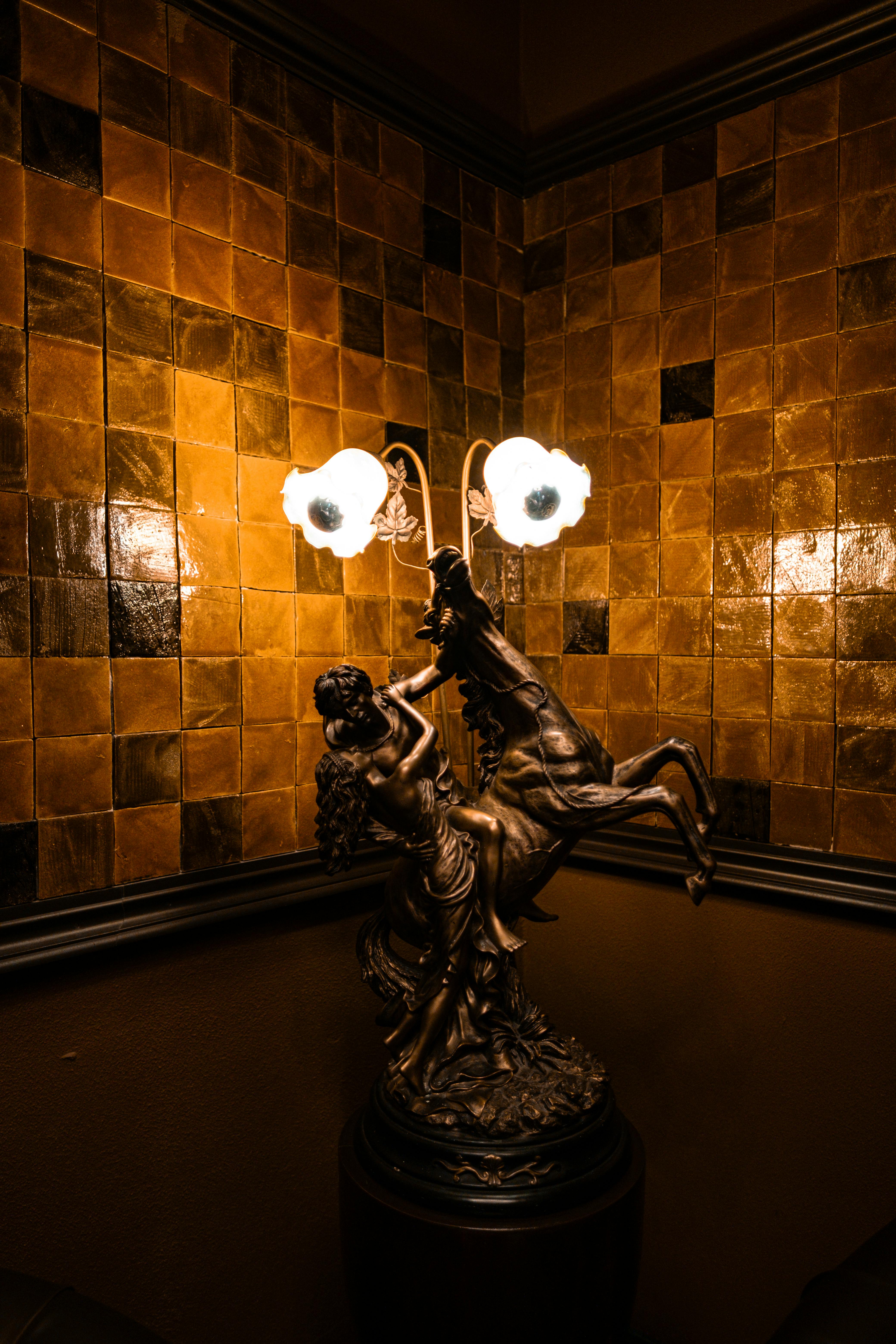 Free Antique lamp with intricate sculpture and glowing lights in a rustic tiled corner, creating a warm ambiance. Stock Photo