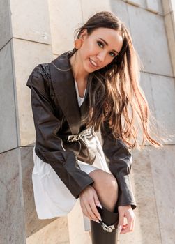 Fashionable young woman in a black leather jacket poses stylishly outdoors against a stone wall.