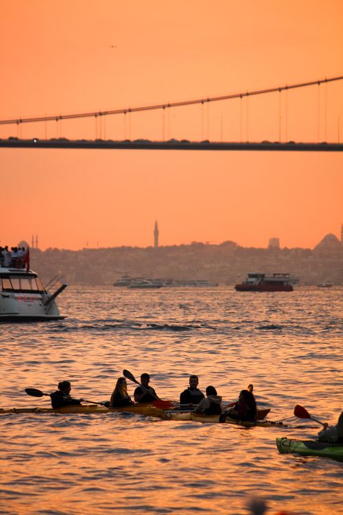Free Kayakers paddle beneath the Bosphorus Bridge at sunset, silhouetted against an orange sky. Stock Photo