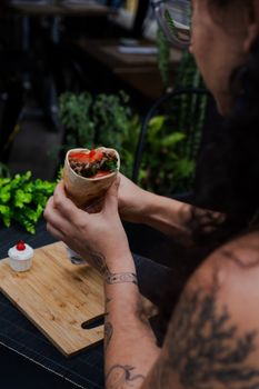 A woman with tattoos holds a fresh vegetable wrap indoors, with plants and a cupcake in the background.