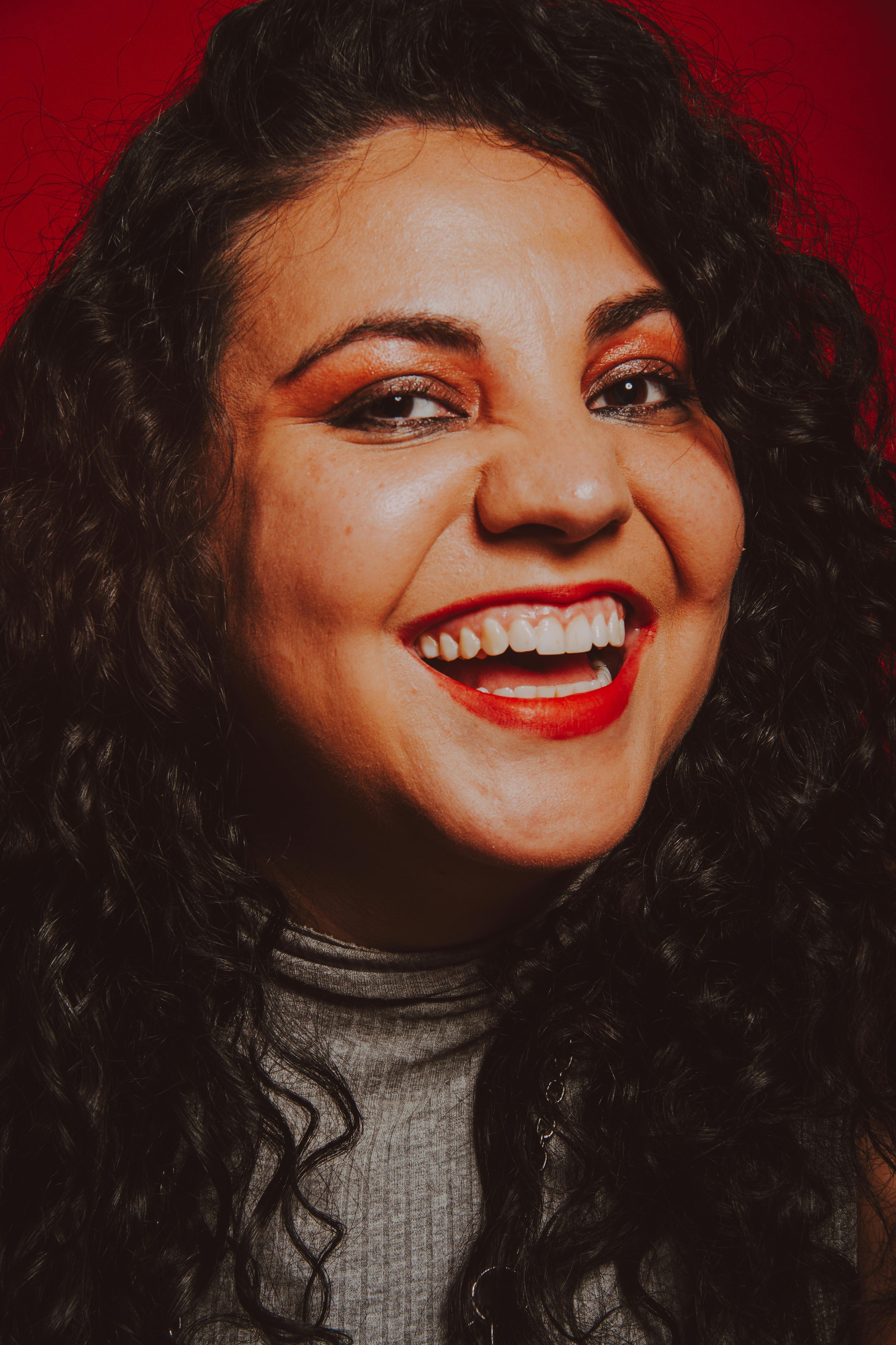 Close-up portrait of a woman with curly hair smiling warmly against a vibrant red background.