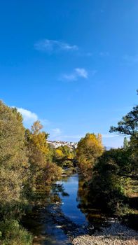 A serene autumn scene with a stone bridge over a calm river, surrounded by colorful trees under a clear blue sky.