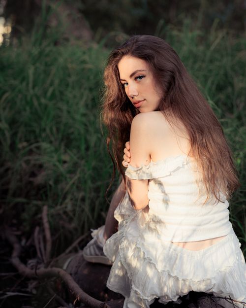 Free Woman in a white dress sitting in a natural outdoor setting, backlit by soft sunlight. Stock Photo