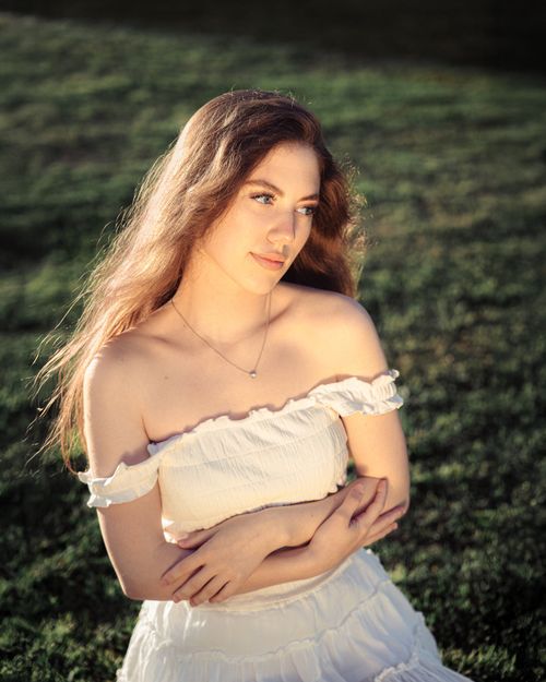 Free Portrait of a young woman in a white dress with long hair sitting on grass, illuminated by soft natural light. Stock Photo
