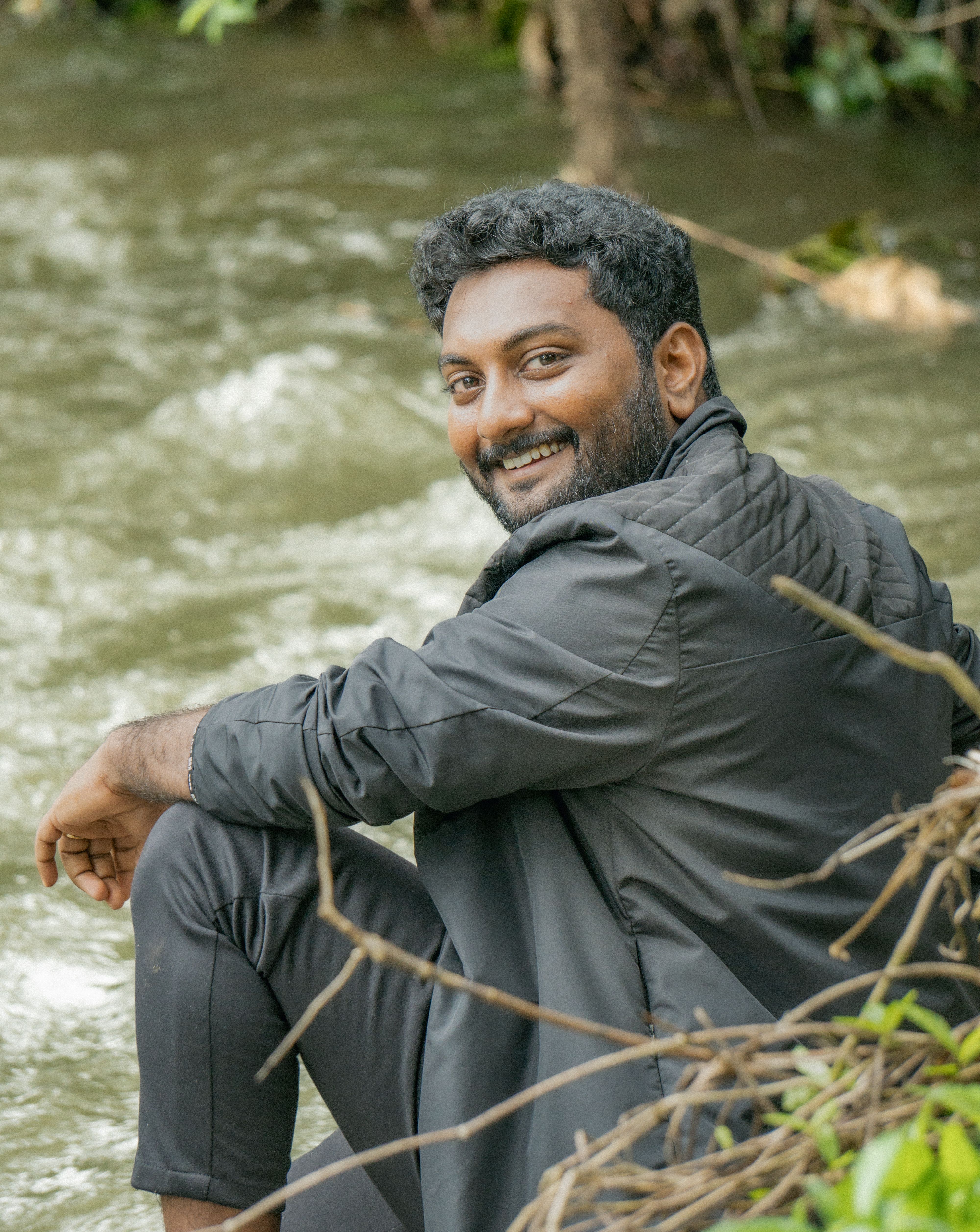 Smiling man in casual attire sitting by a riverbank enjoying nature.