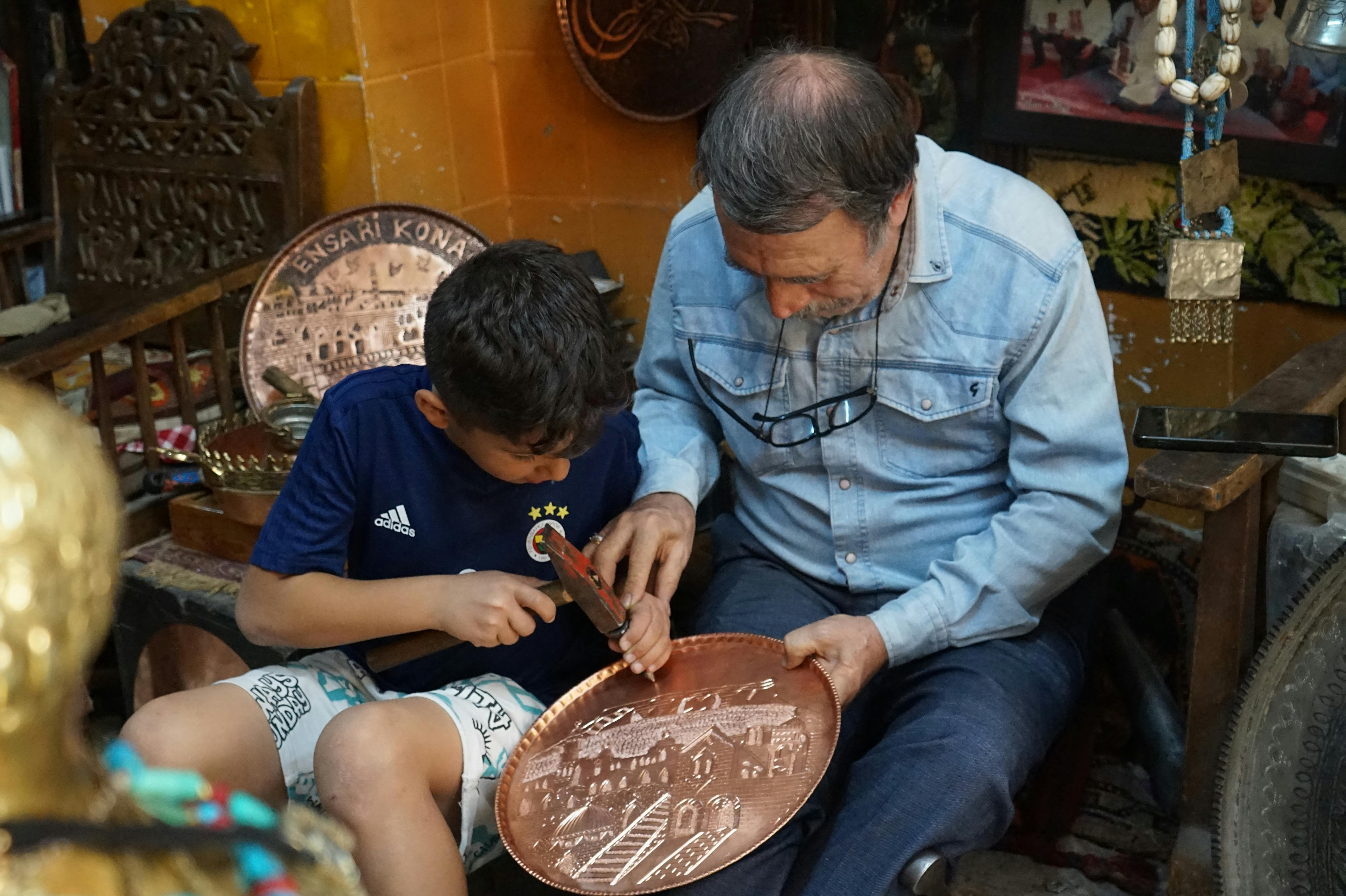 different types of engraving techniques - A child and an elder engraving a copper plate, depicting a traditional craft in a cozy workshop.