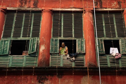 Free A senior woman in traditional attire looks out from a weathered building window with green shutters. Stock Photo