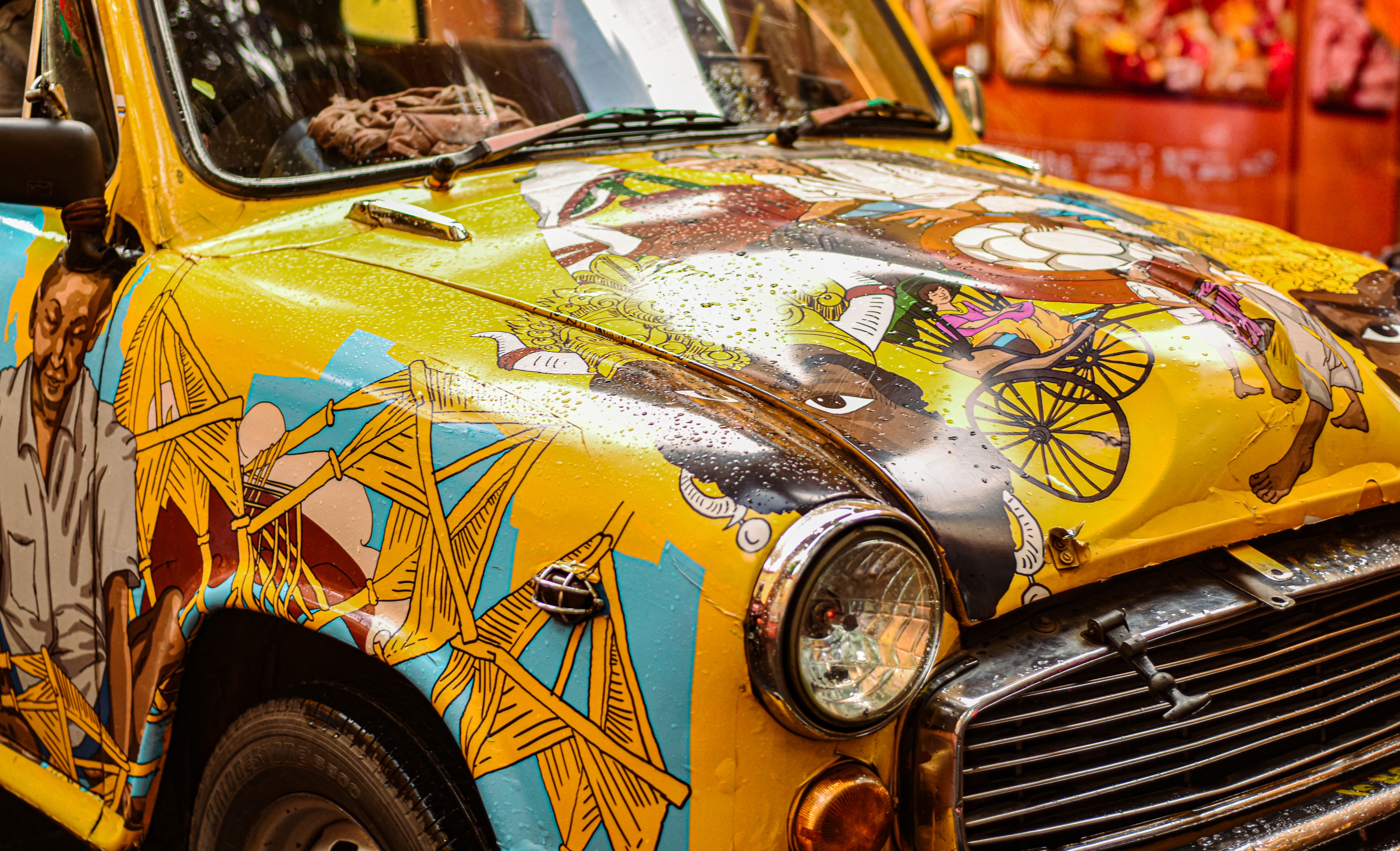 A vibrant vintage car decorated with colorful artistic murals in a sunlit street scene.