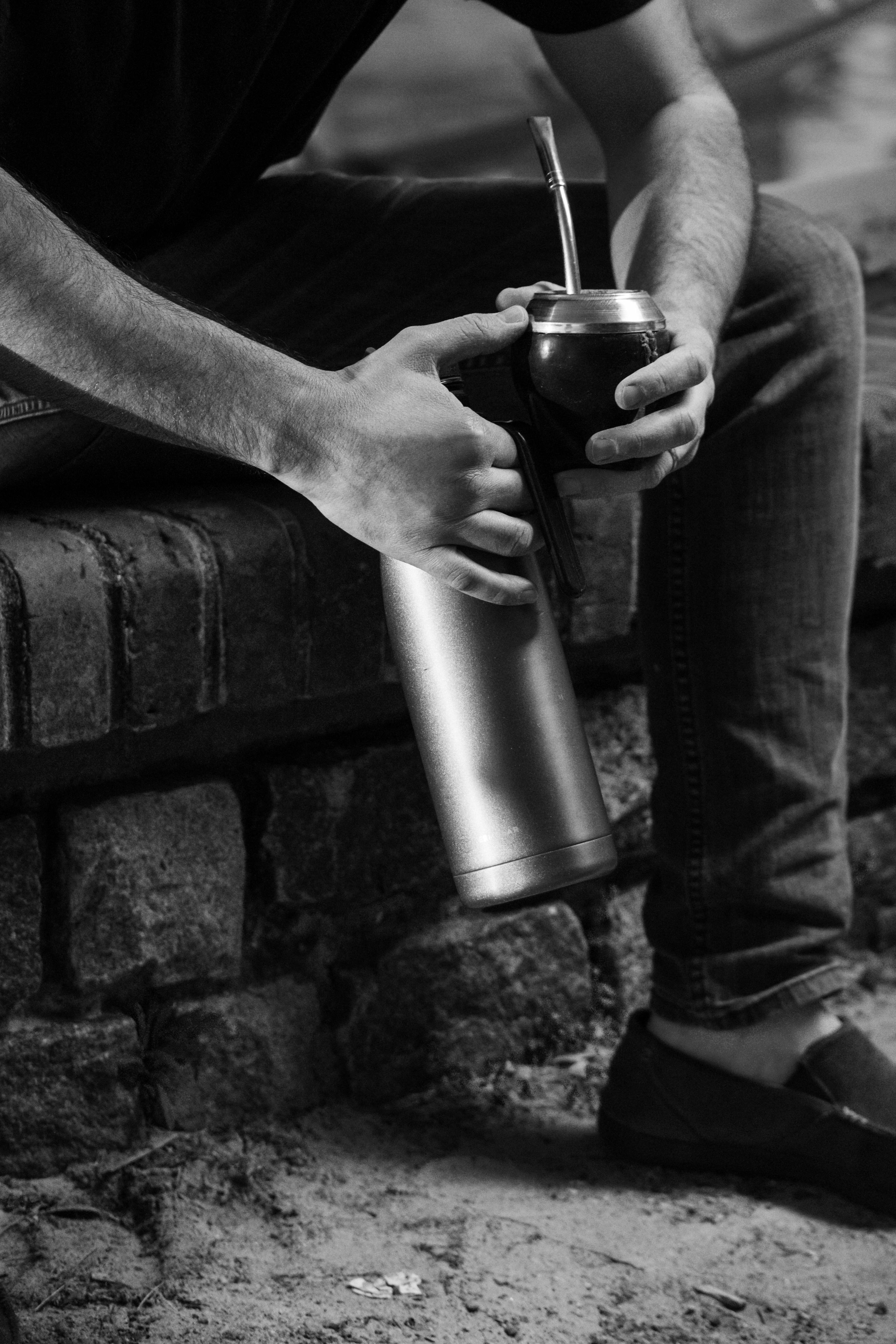 Black and white image of a man drinking mate while sitting on a brick wall outdoors.