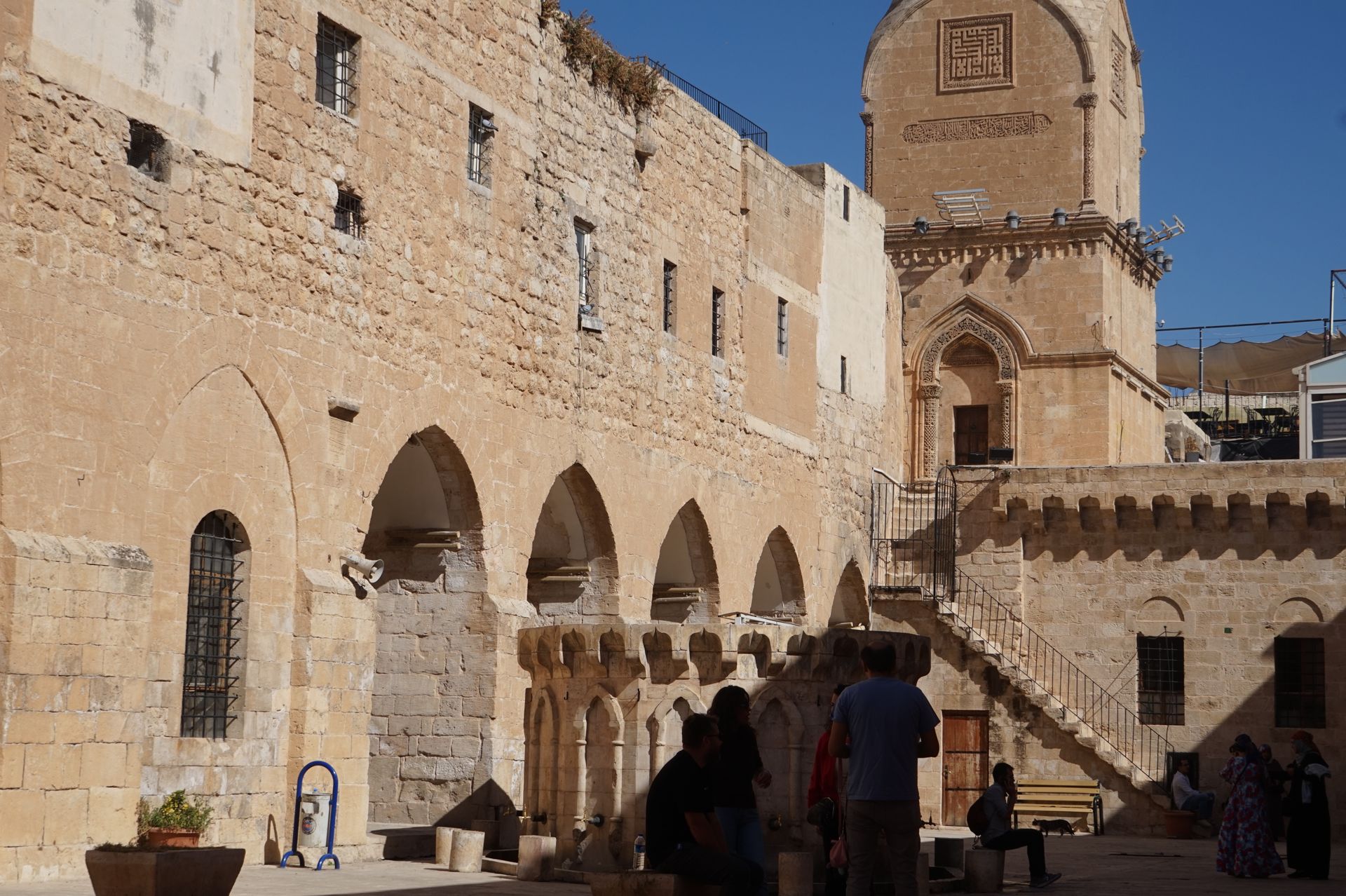 People gathered near historic stone architecture with arches and ancient tower.