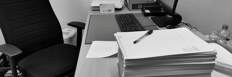 Black and white image of a cluttered office desk with a computer, chair, and paperwork.
