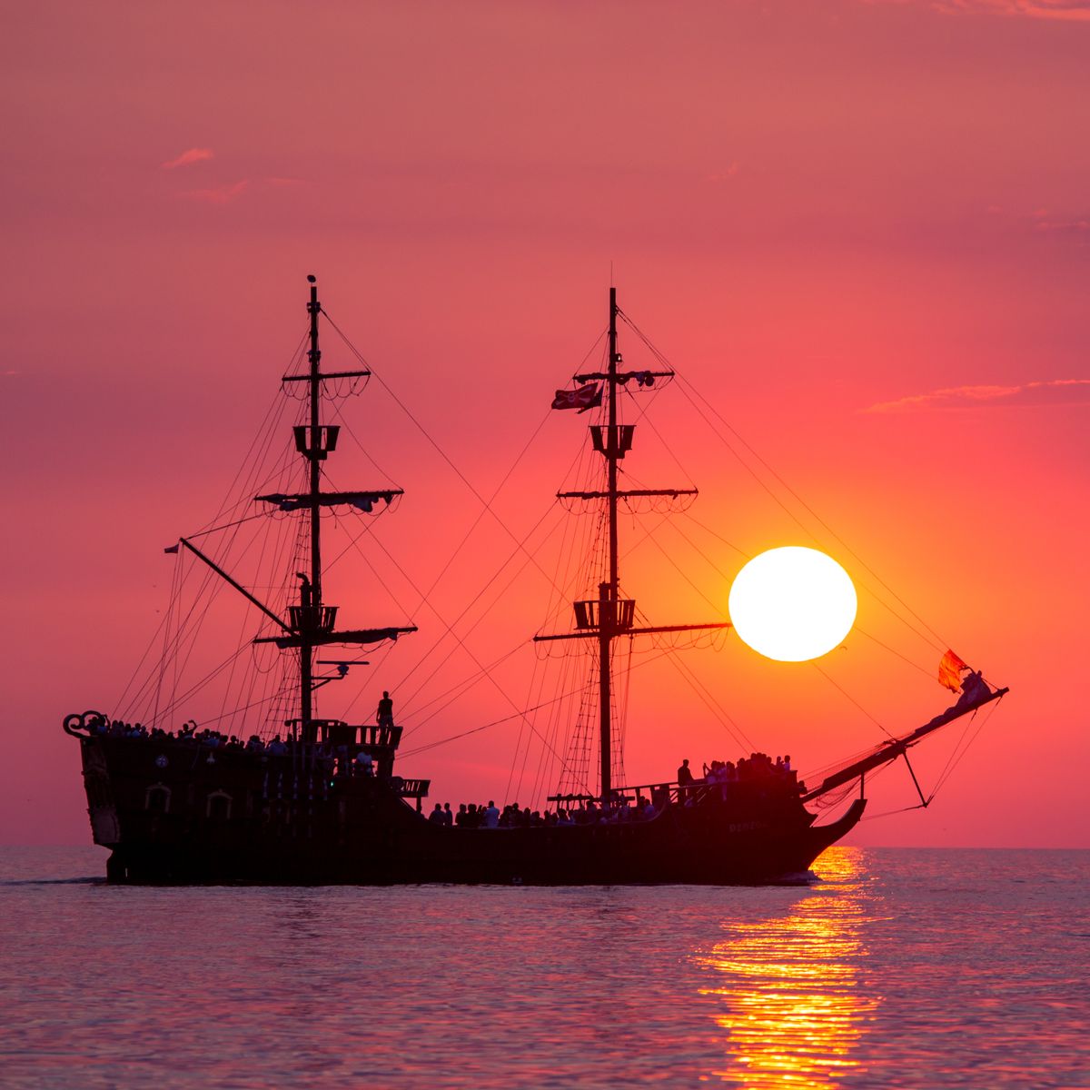 Silhouette of a tall wooden sailing ship against a deep orange sunset horizon, illustrative photo