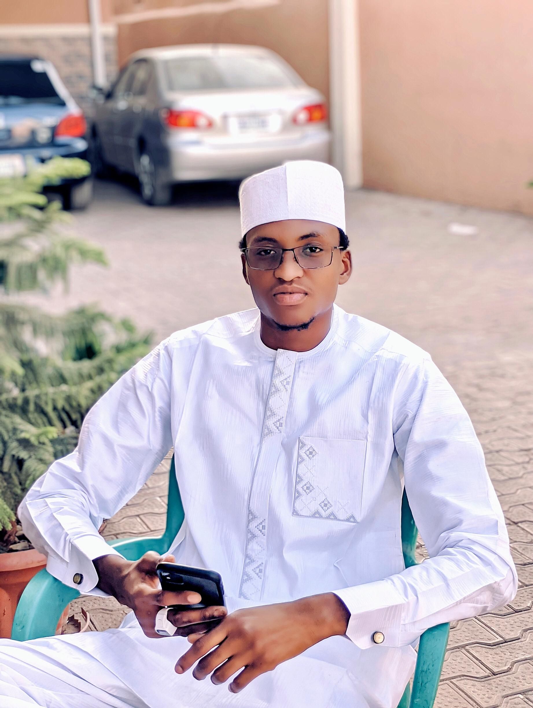 A young man in white traditional clothing sits outdoors holding a smartphone.