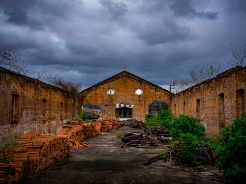 Free Old abandoned warehouse with brick walls and storm clouds above, featuring piles of bricks and overgrown vegetation. Stock Photo