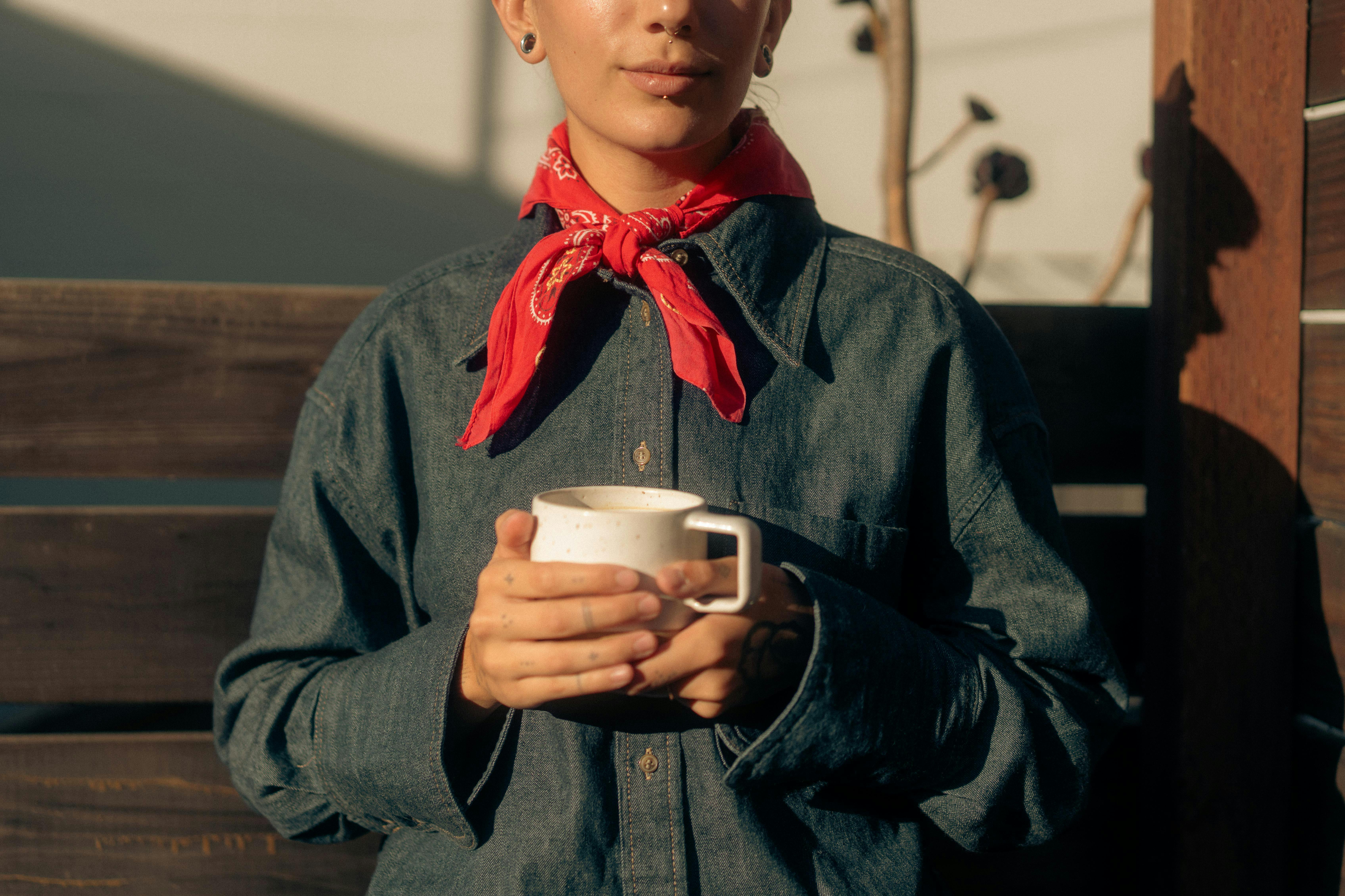 Gratis Un adulto elegante con una bandana rossa e una camicia di jeans tiene in mano una tazza all'aperto, sotto la calda luce del sole. Foto a disposizione