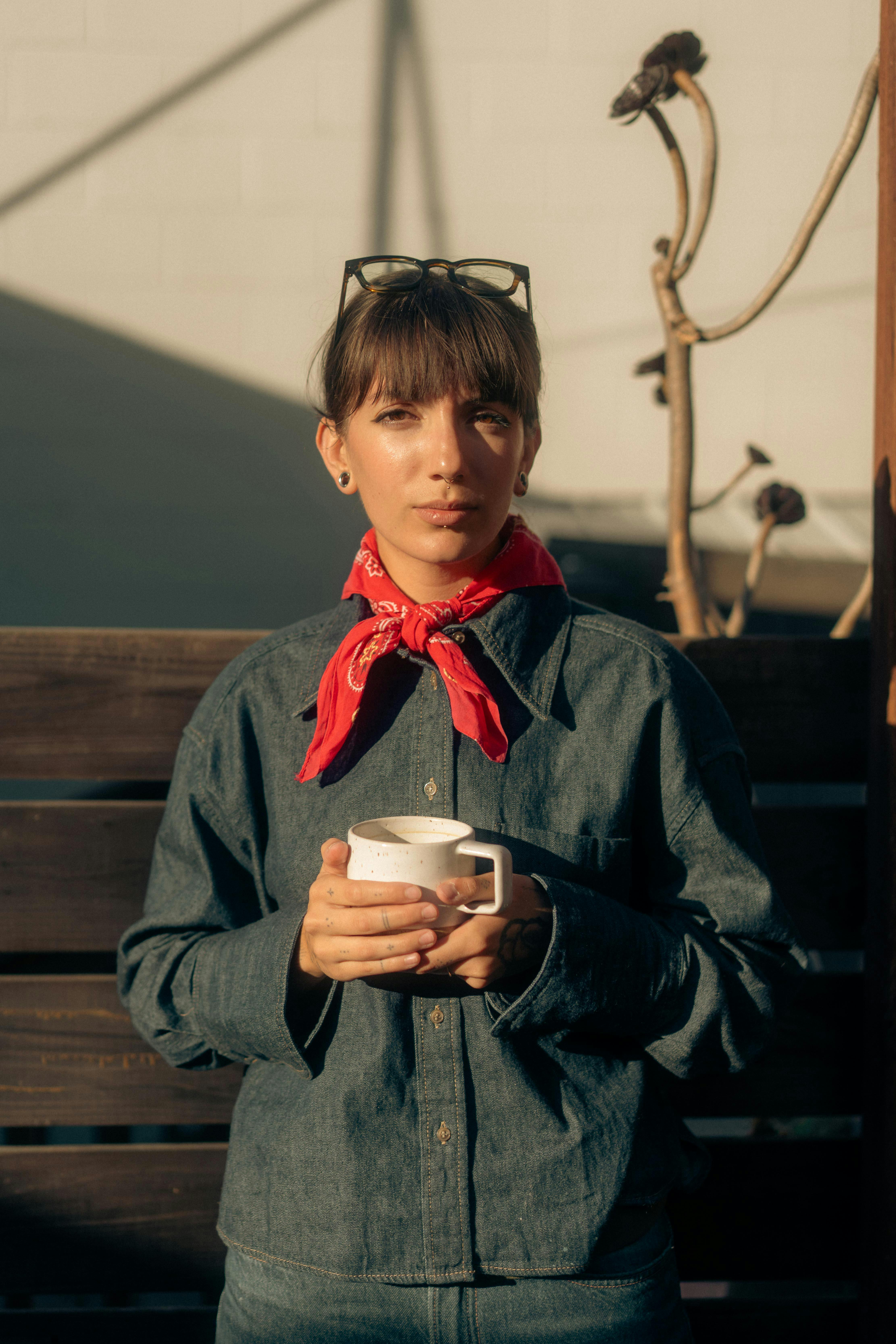 무료 햇볕이 잘 드는 야외에서 빨간 스카프를 두른 데님 차림의 여성이 커피 머그잔을 들고 있는 모습입니다. 스톡 사진