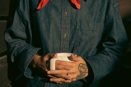 Person holding a coffee cup, wearing denim with tattooed hands in warm light.