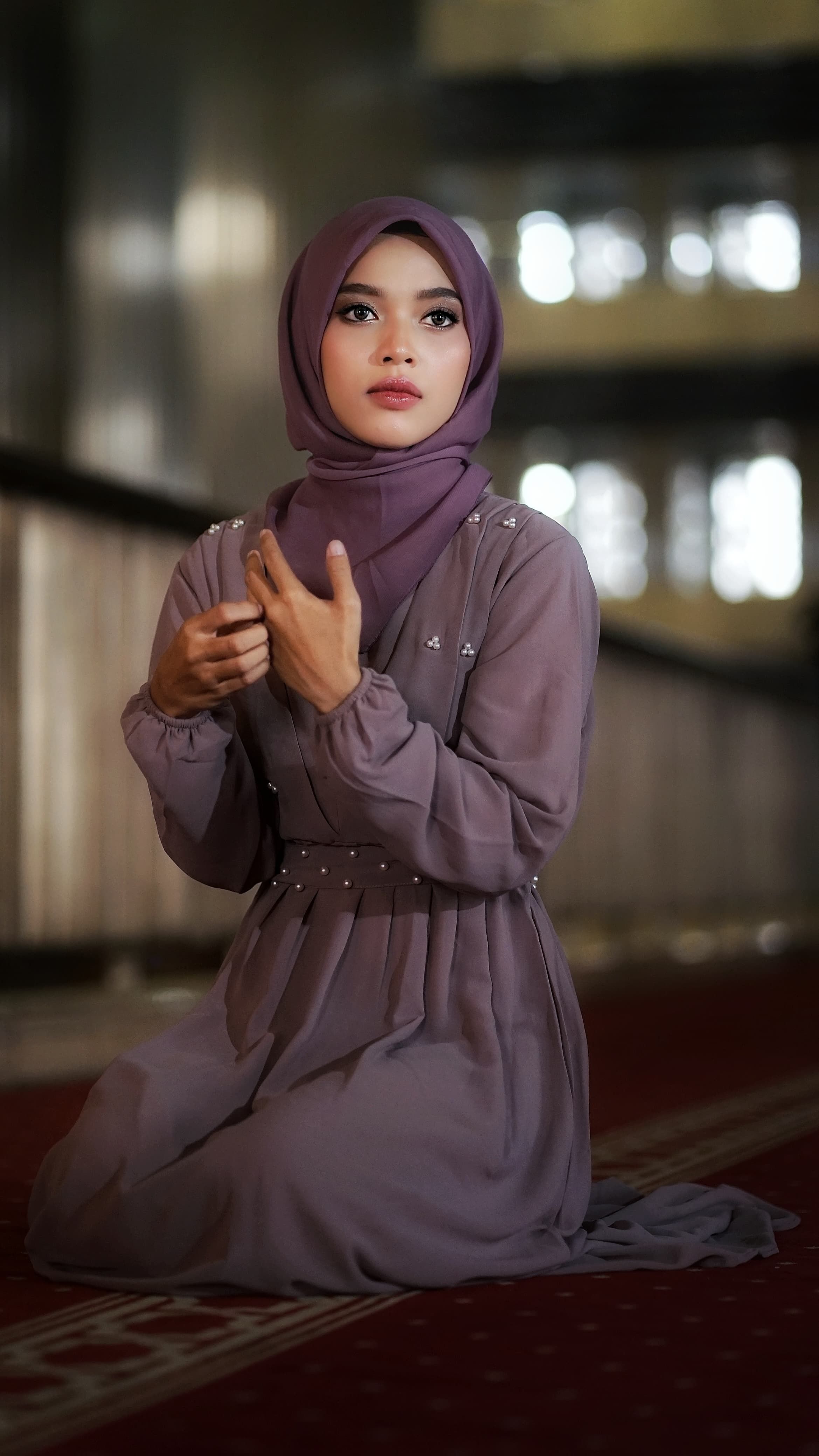 A woman wearing a hijab kneels on a red carpet inside a mosque, reflecting devotion and serenity.