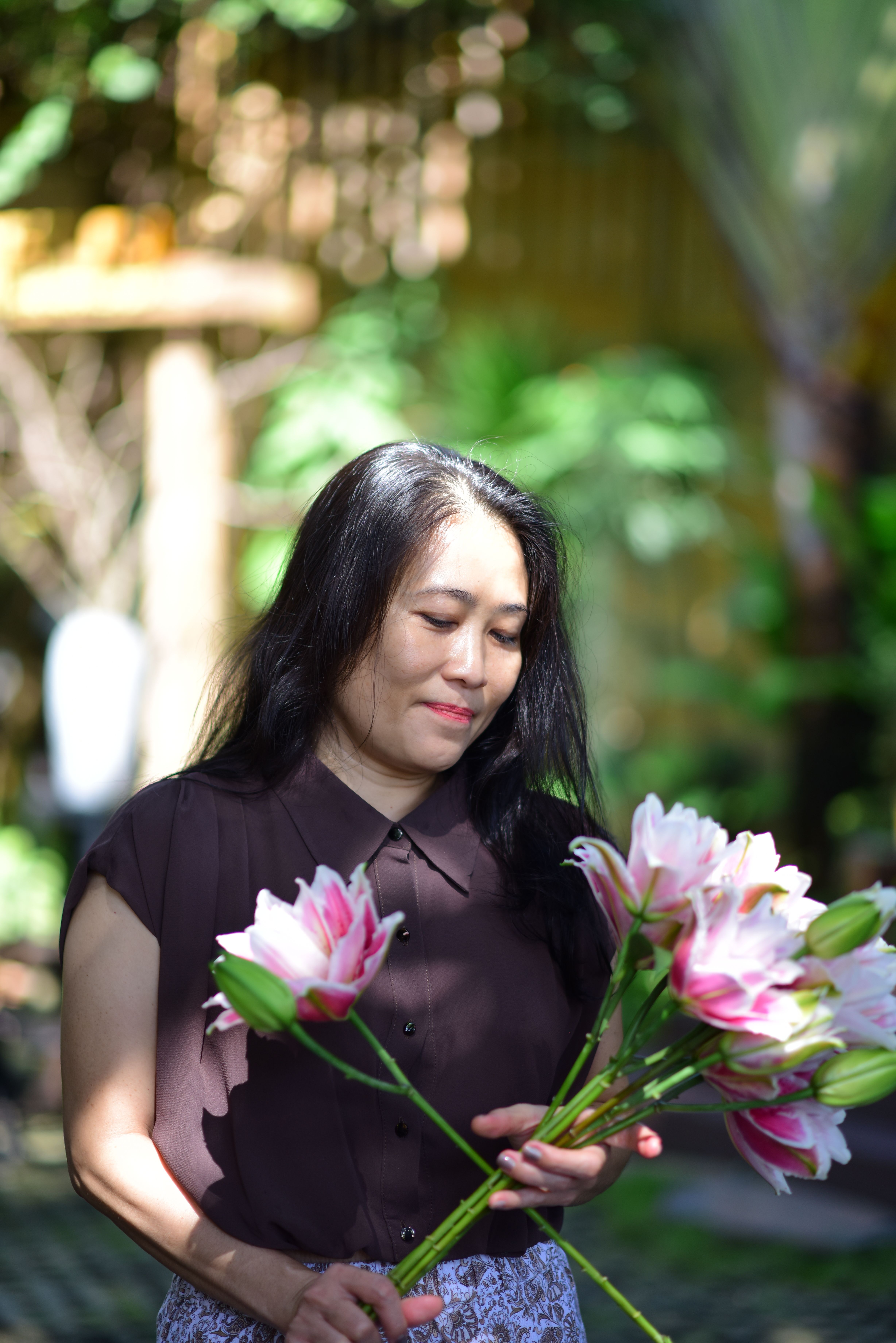 A serene woman holding pink lilies in a sun-dappled outdoor garden with lush greenery.