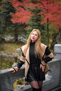 Elegant woman in autumn attire posing on a stone bridge with vibrant fall foliage in the background.