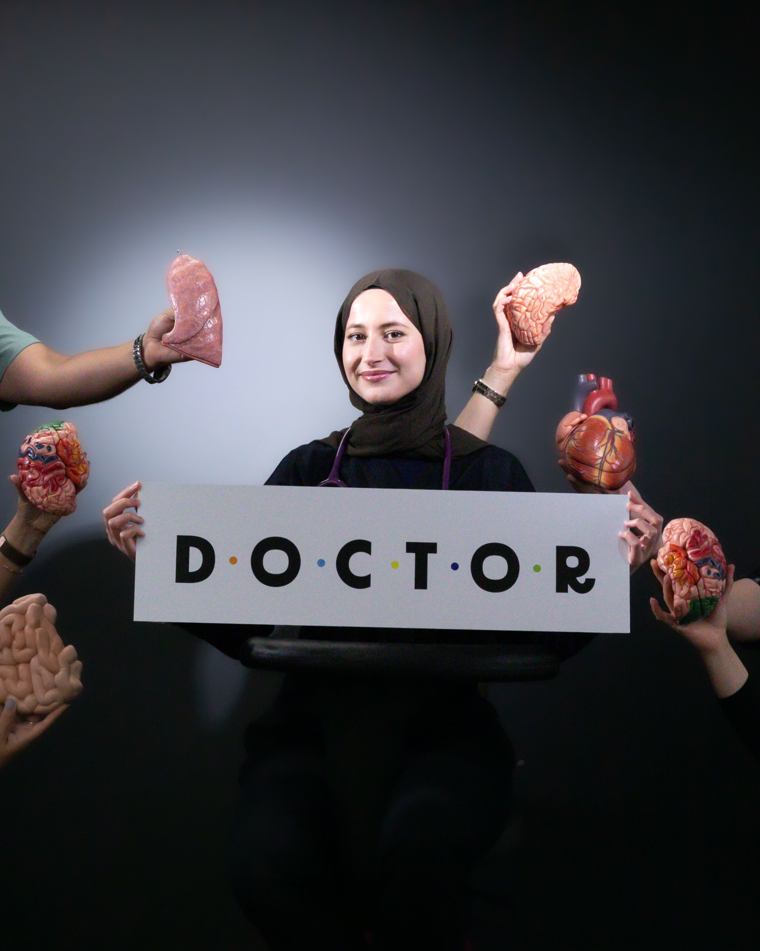 Smiling medical graduate holding a doctor sign surrounded by anatomical models.