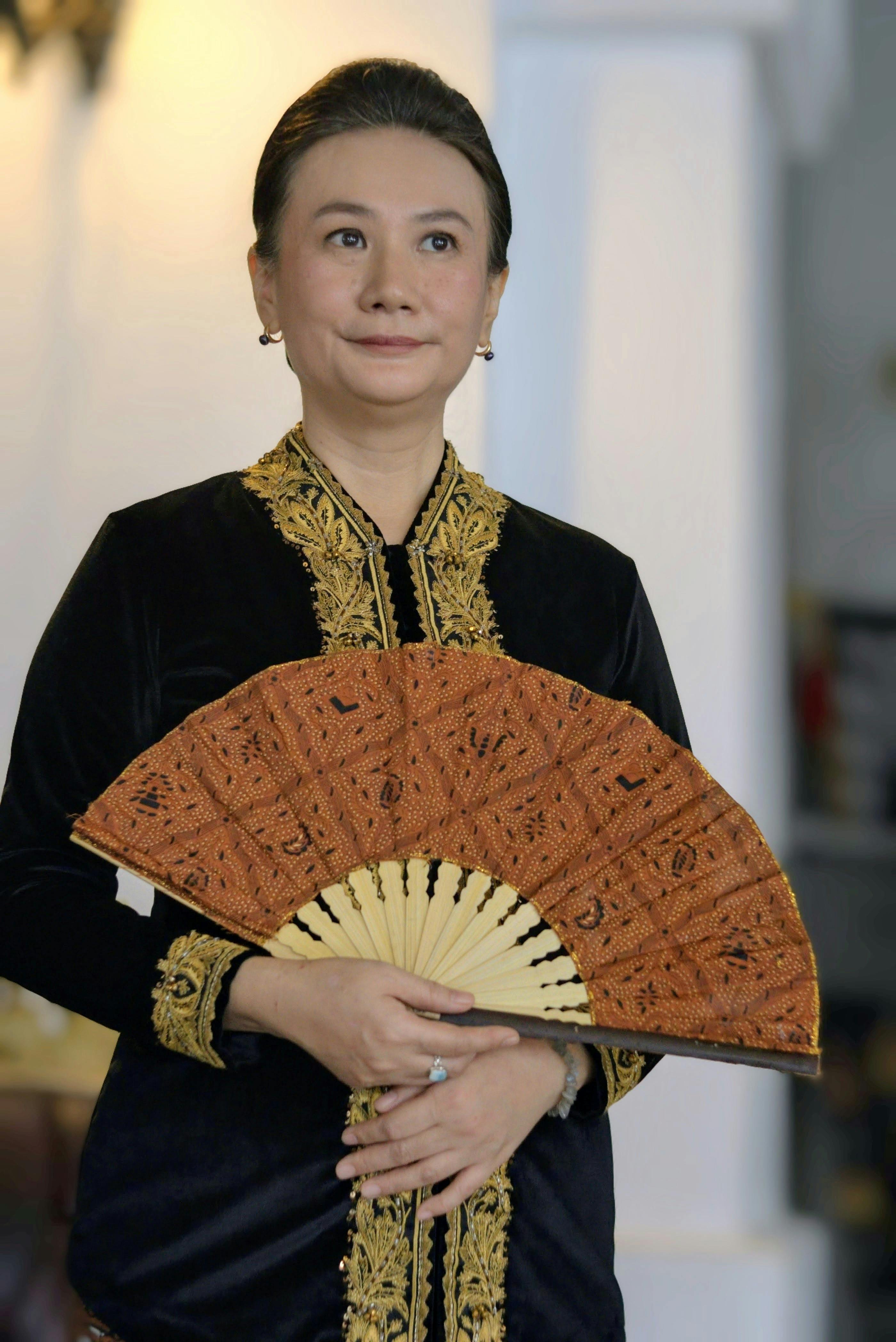 Woman in elegant Indonesian traditional attire holding a batik fan indoors.
