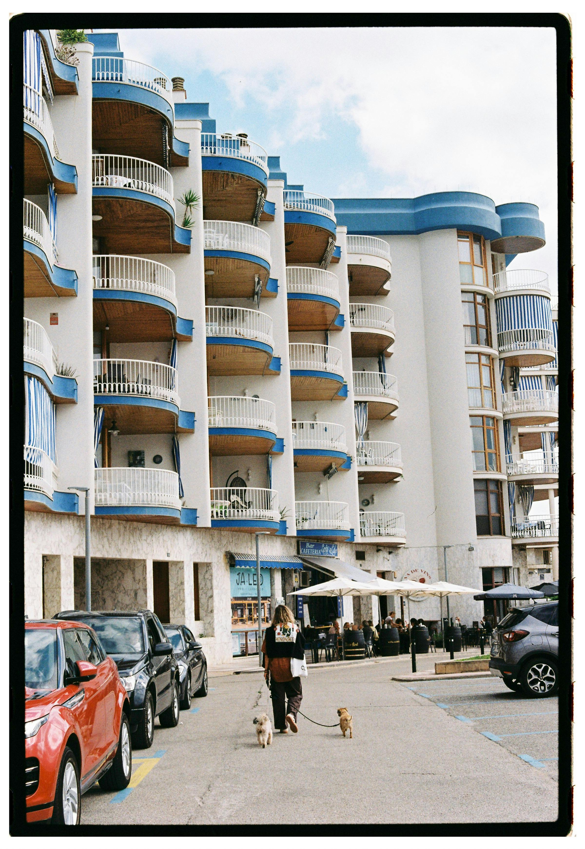 Urban scene of a multi-story hotel with parked cars and people walking dogs.