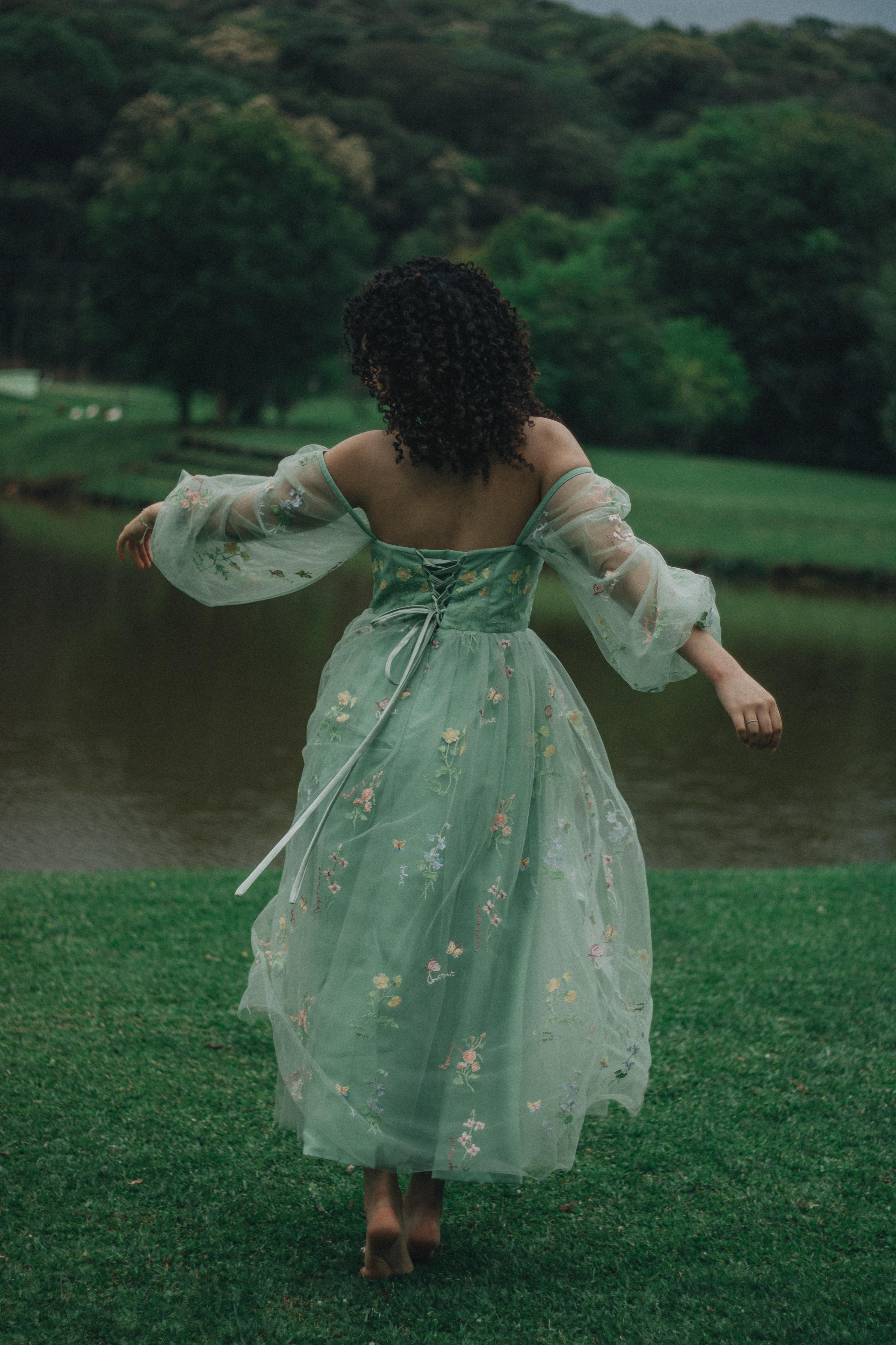 A woman in a flowing green dress gracefully dances near a serene lake surrounded by lush greenery.