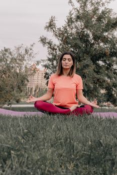 A woman practicing yoga on a grass field in a park, surrounded by trees and buildings.