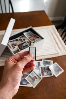 A hand holding a USB drive beside a collection of old printed photos on a table.
