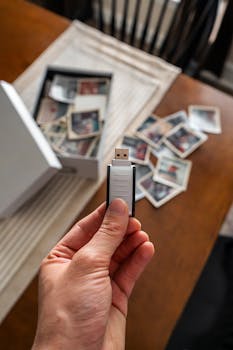 Hand holding USB drive next to scattered vintage photos, symbolizing digital preservation.