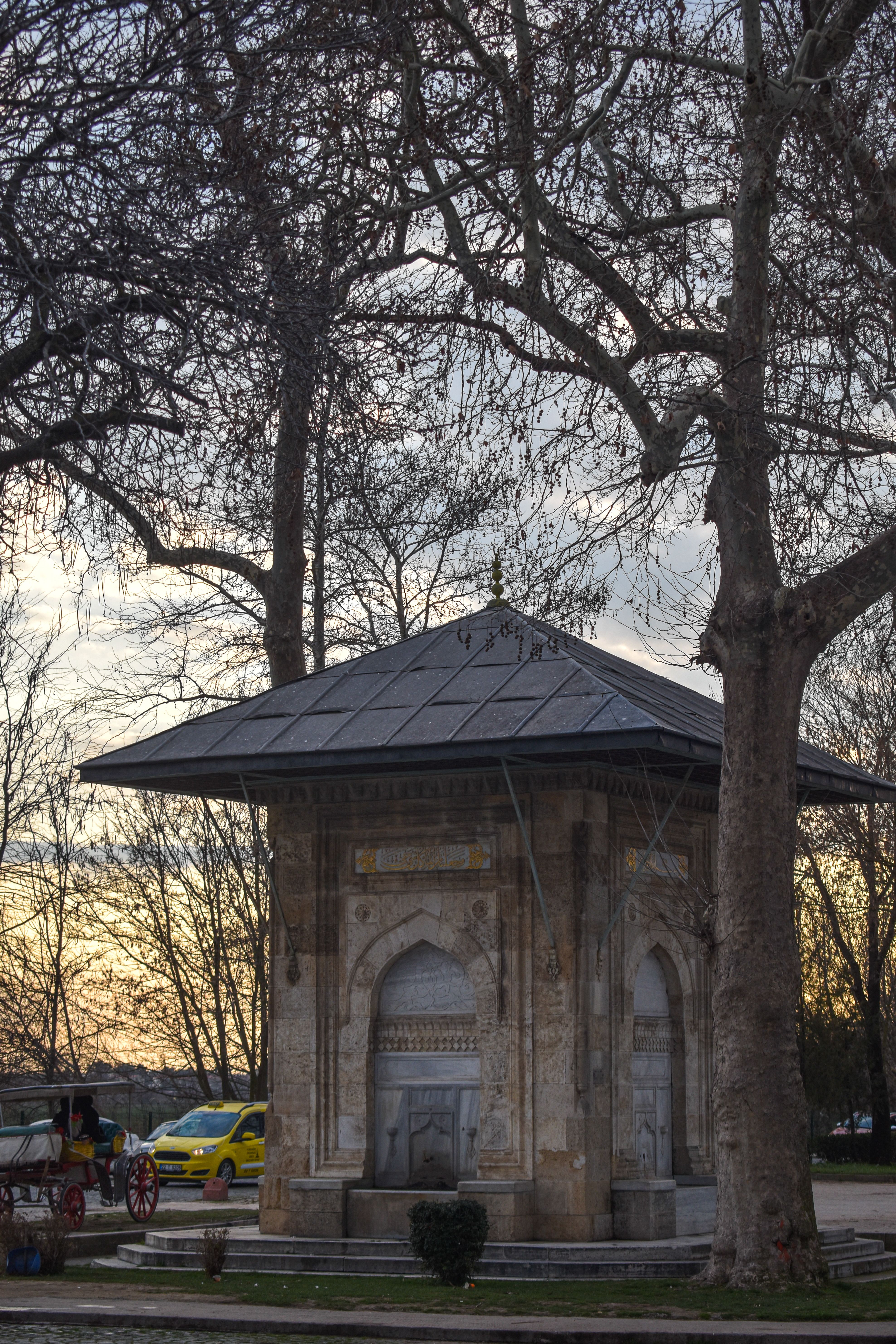 An ancient Ottoman fountain in a park at sunset with bare trees and a peaceful ambiance.
