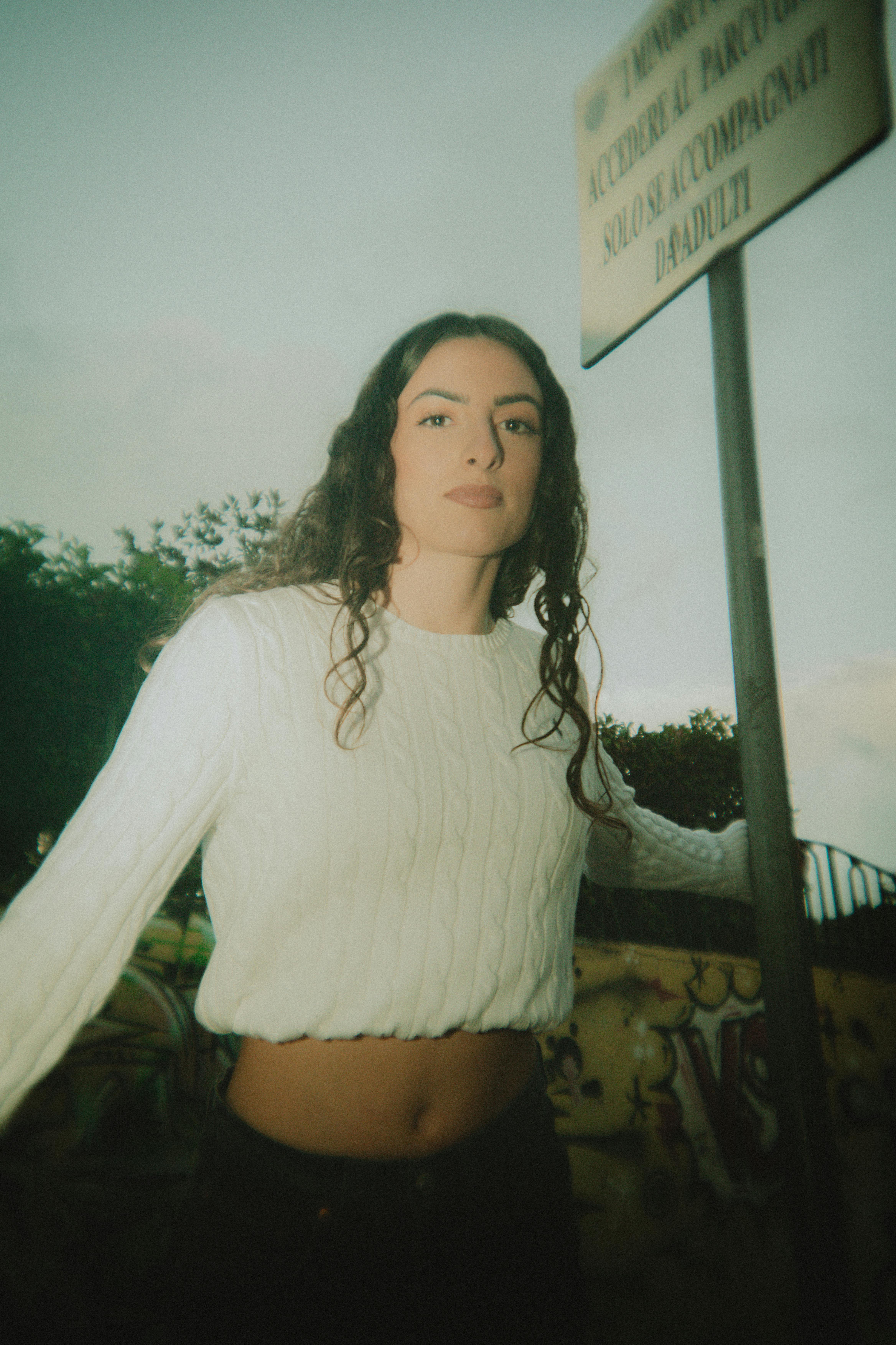 Young woman wearing a white sweater poses near a sign outdoors with a soft, nostalgic feel.