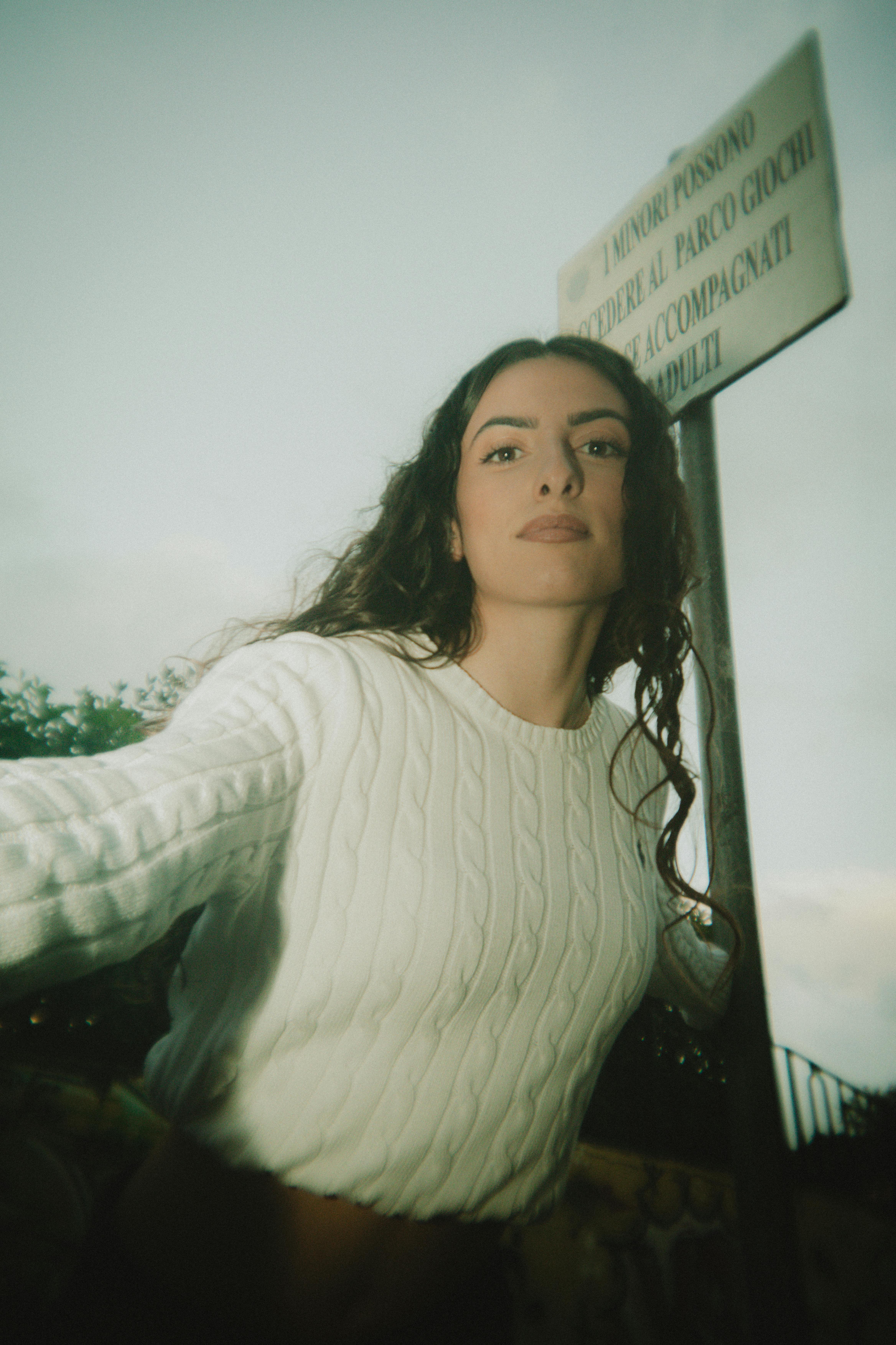 Young woman outdoors in a white sweater standing near a park sign during the day.