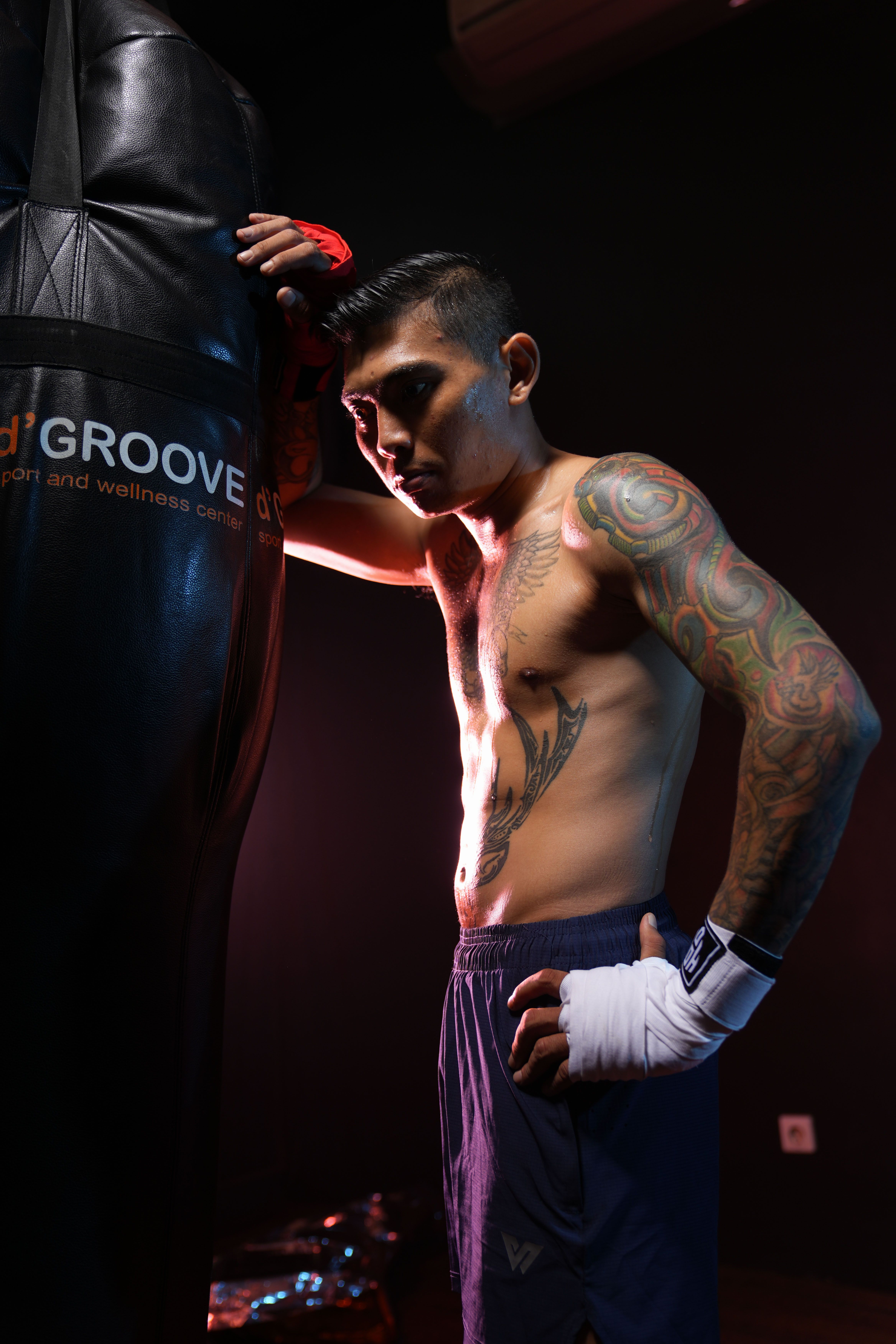 Focused boxer with arm tattoos and wrapped hands standing by punching bag in dimly lit gym.