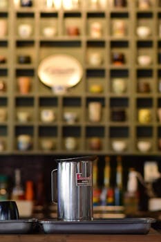 Stainless steel coffee pitcher on a tray in a cozy cafe with a decorative shelf background.