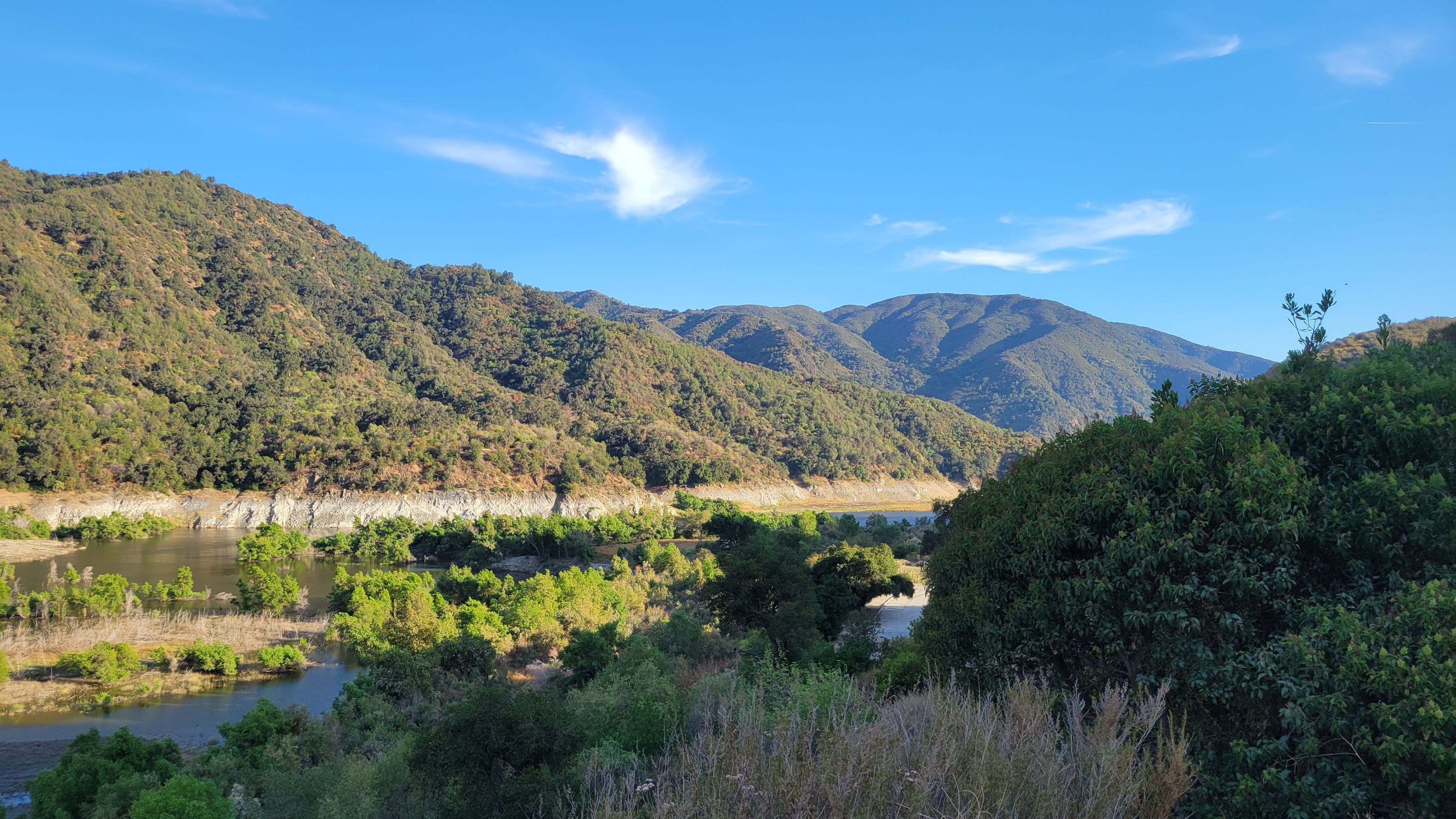 Peaceful mountain view with lush green vegetation and a flowing river under clear blue skies.